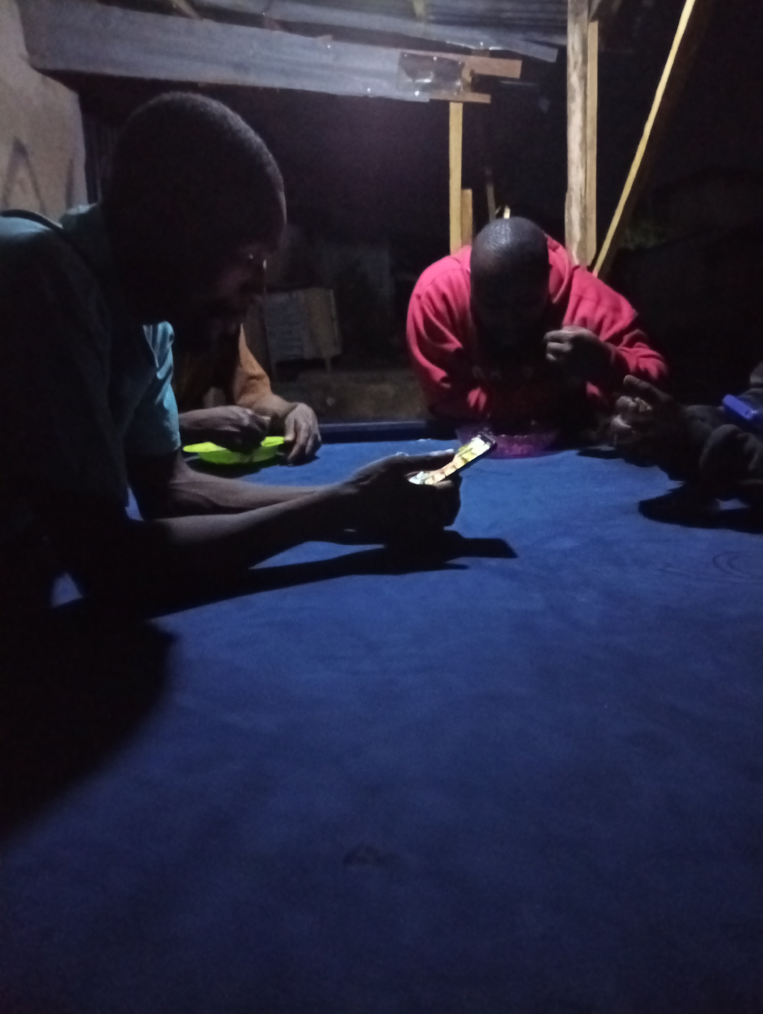 Four individuals are gathered around a blue-topped table in an indoor setting. One person is holding and looking at a smartphone. Another person is eating from a pink bowl. The setting appears to be dimly lit, with a corrugated metal roof and wooden support beams. The exact location in Jos, Nigeria, cannot be determined from the image.