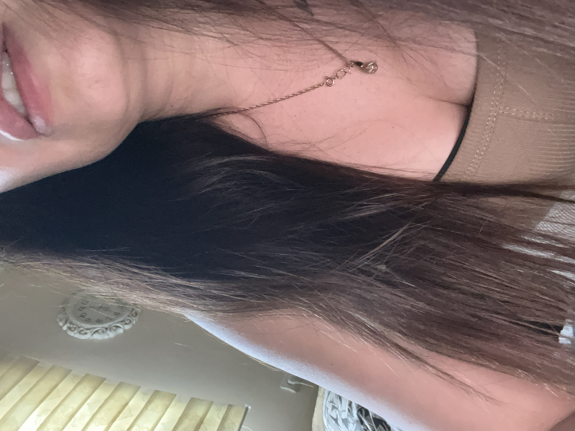 A medium shot photograph shows a woman's face and upper body. The woman has long, dark brown hair that is falling around her face and shoulders. She is wearing a tan tank top and a delicate gold necklace with a small pendant. Her lips are parted slightly, revealing her teeth. To the left of the woman's head, a white decorative clock is mounted on the wall. Behind her, a window with vertical blinds is visible. The scene appears to be indoors, possibly in a bedroom, with soft lighting.