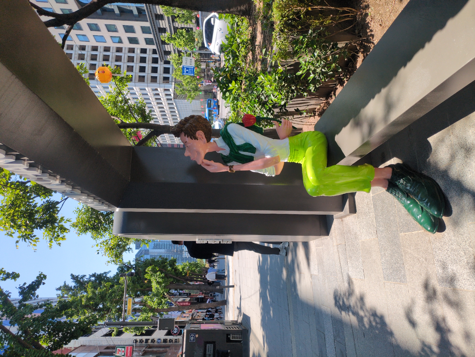 The image shows a brightly colored statue of a man seemingly peering down from a ledge along a city sidewalk in Seoul, South Korea. The statue is notable for its vibrant green pants and shoes, a white shirt with a green scarf, and a stylized facial expression. The statue is mounted on a grey ledge or architectural feature, under which pedestrians walk along the sidewalk. Behind the statue and ledge, there's a green hedge and then a city street with cars and a tall building with a grid-like window pattern. Along the sidewalk in the foreground are various shops and buildings with Korean signage, including a 7-Eleven store and a CCTV warning sign. The overall scene suggests a public art installation in an urban setting.