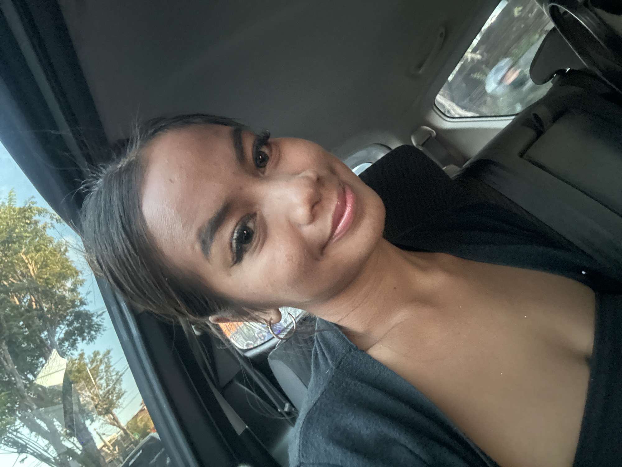 A woman is taking a selfie inside a car. She is looking at the camera with a slight smile. She is wearing a dark top and a grey cardigan. There is a circular earring in her left ear. The car appears to be parked on a street in North Kuta, Indonesia, as indicated by the presence of trees and buildings visible through the car windows. The sun is setting, casting a warm glow on the scene.