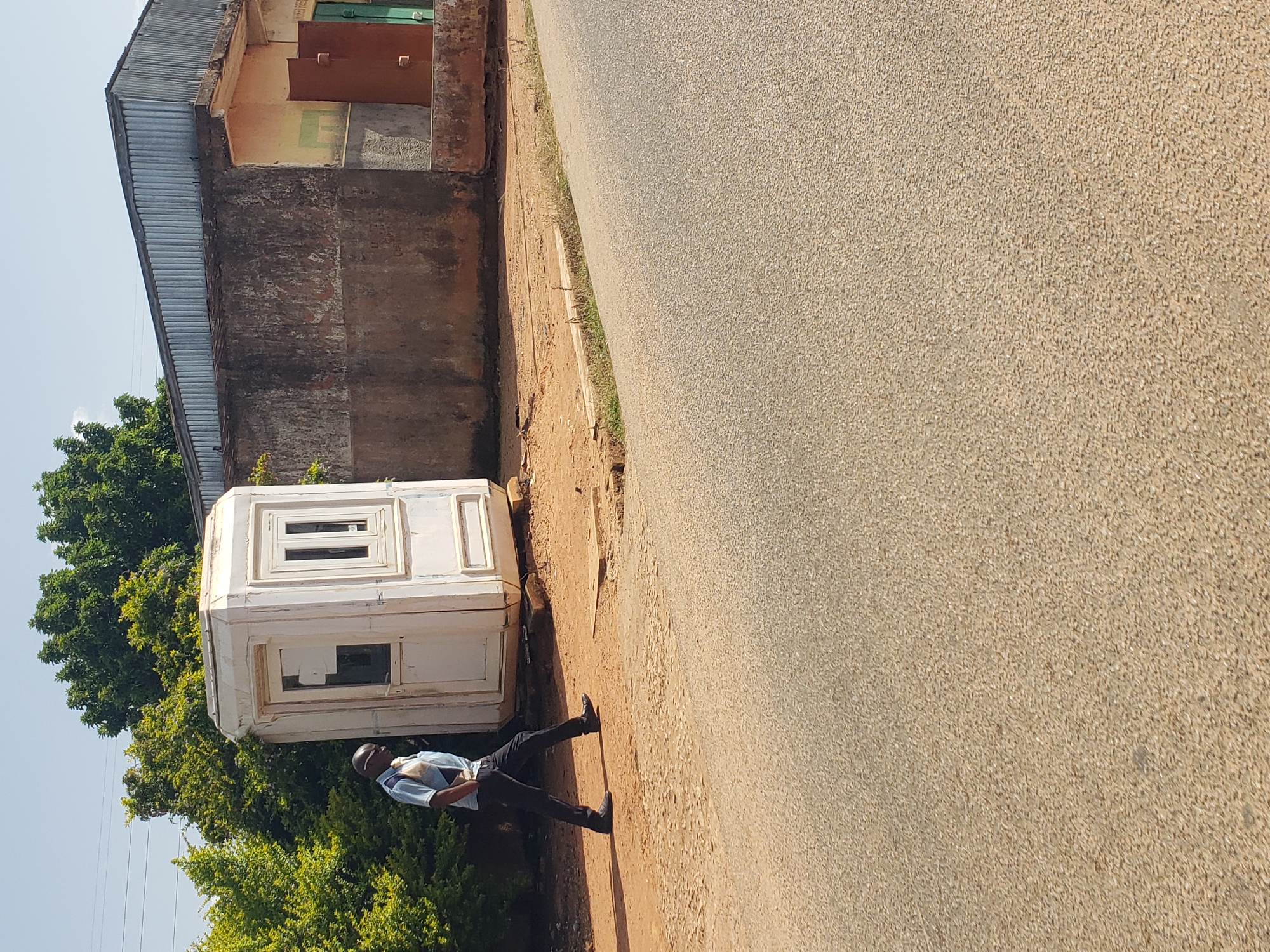 A bald African man wearing a light blue polo shirt and dark trousers walks from left to right in front of a white octagonal security booth. The booth has two windows on its visible side, one rectangular and one square, both with white frames. Behind the booth, a corrugated metal roof extends over a building with rough concrete walls. To the left of the booth, lush green trees and bushes are visible. The ground is a mix of reddish-brown soil and gravel, with a paved road in the foreground. The scene appears to be an outdoor area in Gura Top, Nigeria, under a clear, bright sky.