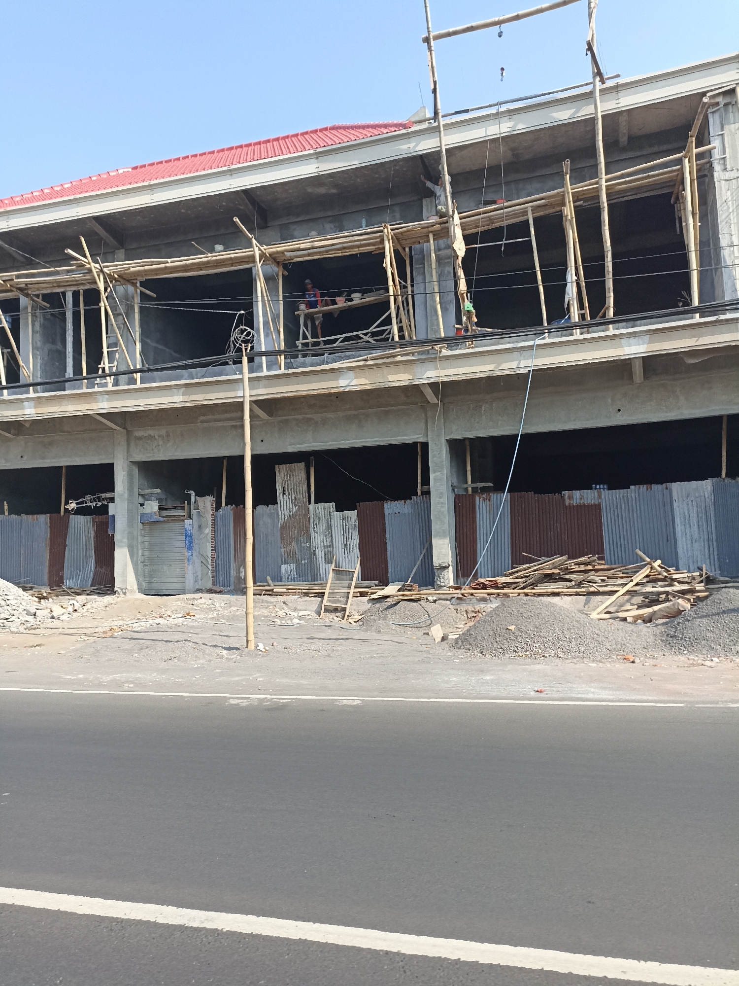 A construction site is depicted in Lumajang, Indonesia. The main subject is a multi-story building under construction, with concrete floors and walls, and a red tiled roof. Bamboo scaffolding is erected around the upper floors. Several workers are visible on the scaffolding, engaged in construction activities. The ground level is a dirt and gravel area, with piles of construction materials such as gravel and lumber. Corrugated metal sheets are used as temporary barriers. A paved road with white lane markings runs in the foreground, indicating the site's proximity to a street. The sky is clear and blue.