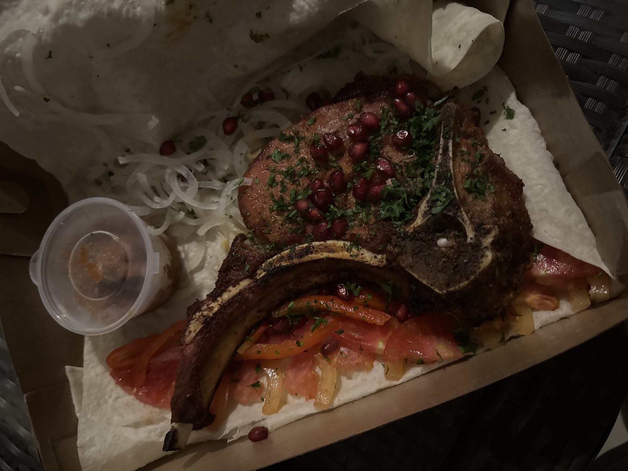 A close-up shot displays a substantial meal presented in a cardboard takeout box. The central focus is on two large, grilled meat chops, likely pork or lamb, with prominent bones. The meat has a dark, seared exterior and is generously garnished with bright red pomegranate seeds and finely chopped fresh green herbs, possibly parsley or cilantro.
Beneath and around the meat, several accompanying ingredients are arranged. Thinly sliced white onions are visible, particularly on the left side of the box. Sliced red tomatoes and other lighter-colored sliced vegetables, possibly bell peppers, are positioned under one of the meat pieces. The entire ensemble rests on and is partially wrapped by soft, white flatbread, resembling lavash or pita, which lines the bottom and sides of the box.
To the left, a small, clear plastic container with a lid holds a reddish-brown sauce. The overall lighting is dim, suggesting the photo was taken in the evening or at night, possibly under artificial light. The background features a dark, textured surface, possibly a wicker or rattan table, hinting at an outdoor or casual setting. The meal appears ready for consumption. No text is visible in the image.