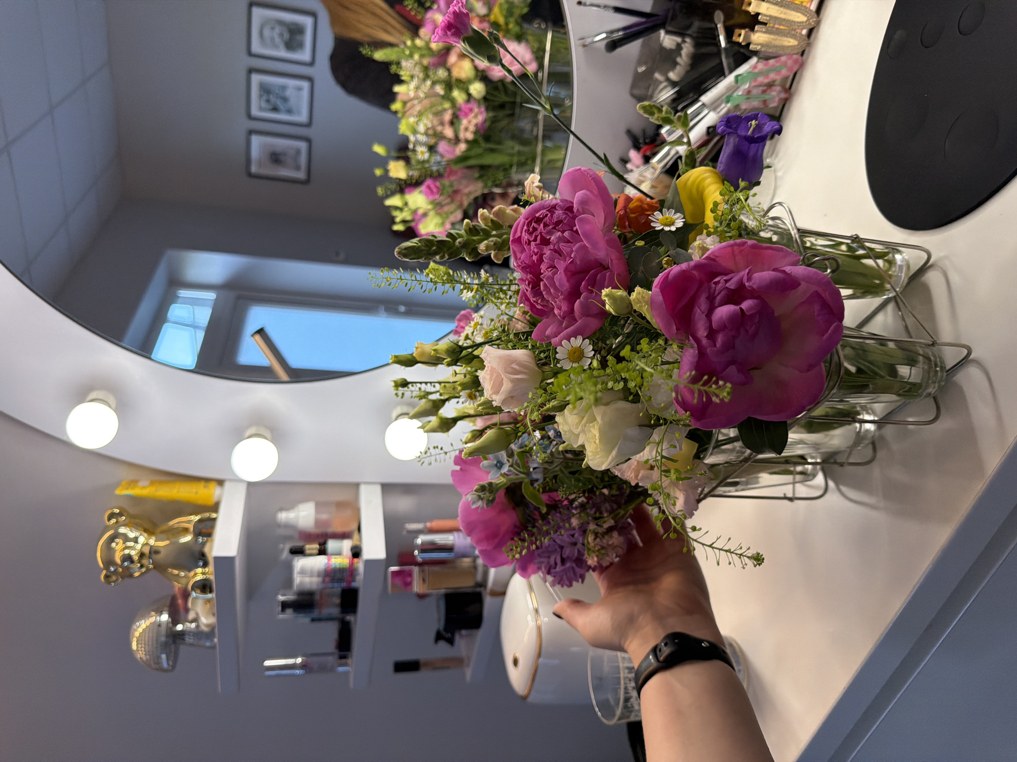 This image captures an indoor scene at a vanity area, where a person is holding a vibrant bouquet of fresh flowers. The perspective is slightly tilted, with the person's hand entering the frame from the lower right, holding the flowers towards the center.
The bouquet is rich with color and variety, prominently featuring large pink peonies, several smaller white daisies, light pink carnations, some deep purple bell-shaped flowers, and various green foliage and smaller white blossoms. The hand holding the bouquet has a black wristwatch on the wrist.
To the left of the bouquet is a large, round mirror, framed with several exposed, glowing light bulbs, illuminating the area. The mirror reflects the room behind the camera, showing a plain light-colored wall with three framed black-and-white pictures hanging vertically. A window, letting in natural daylight, is also visible in the mirror's reflection, suggesting it is daytime. A faint reflection of a person's hair or head can be seen in the upper part of the mirror.
The flowers are held above a white vanity table, which occupies the right side of the frame. On this table, various cosmetic products, bottles, and containers are neatly arranged on white tiered shelves or within open drawers. A golden bear-shaped figurine and another metallic, possibly silver, object are discernible on a shelf at the lower left part of the vanity. A black, circular mat or object with indentations is partially visible on the far right of the table.
The scene suggests a calm, personal space, likely a bedroom or dressing room, well-lit by both the vanity lights and natural light. There is no visible text in the image. The implied activity is either admiring, arranging, or receiving the flowers. While Riga, Latvia is given as context, no visual cues within the image specifically indicate this location.