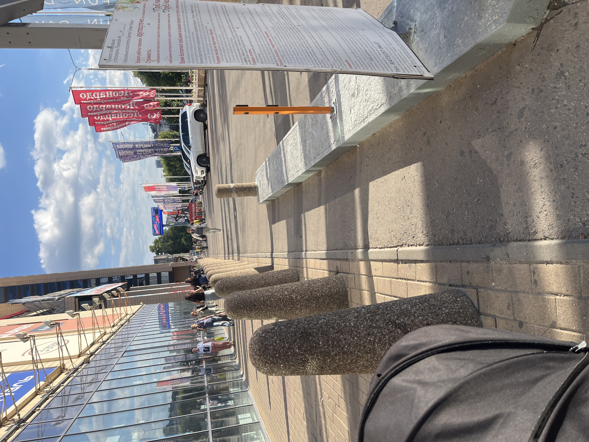 In Voronezh, Russia, the exterior of a modern building with a glass facade is captured on a sunny day with scattered clouds. A paved walkway runs alongside the building, lined with cylindrical concrete bollards. Across the walkway, a row of red and blue flags with Cyrillic text are displayed, flapping in the breeze. Parked cars, including a white SUV, are visible behind the bollards. A large informational sign with text in Russian stands on the right side of the frame. Several people are walking on the sidewalk, some carrying shopping bags. The ground surface transitions from pavement to asphalt, with a concrete curb separating the two. Shadows from the building and bollards are cast across the walkway, indicating the direction of the sun.