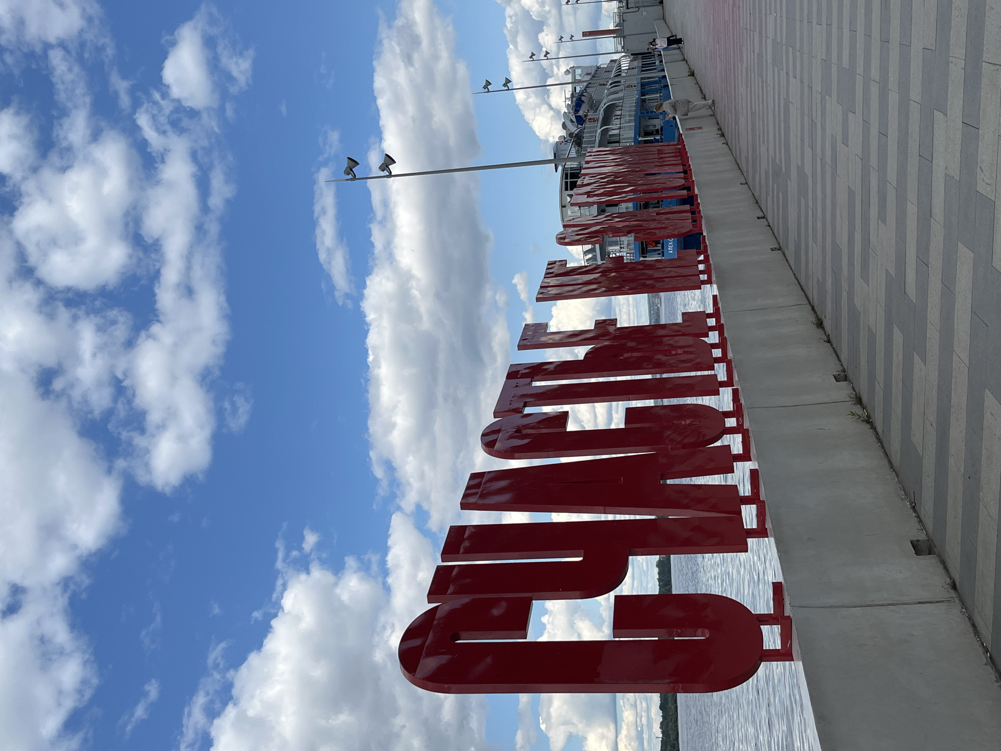 The image depicts a sunny day at the Perm city embankment in Russia. In the foreground, large, red, three-dimensional Cyrillic letters spell out "СЧАСТЬЕ" (meaning "happiness" in Russian). These letters are mounted on a low concrete wall along the embankment. Behind the letters, a wide river is visible, with a blue and white passenger ship docked to the right. The ship has the name "АЛЕКСАНДР" visible on its side. A paved walkway runs alongside the embankment, with a few people strolling. Tall lamp posts with multiple lights are spaced along the walkway. The sky is a vibrant blue with fluffy white clouds.