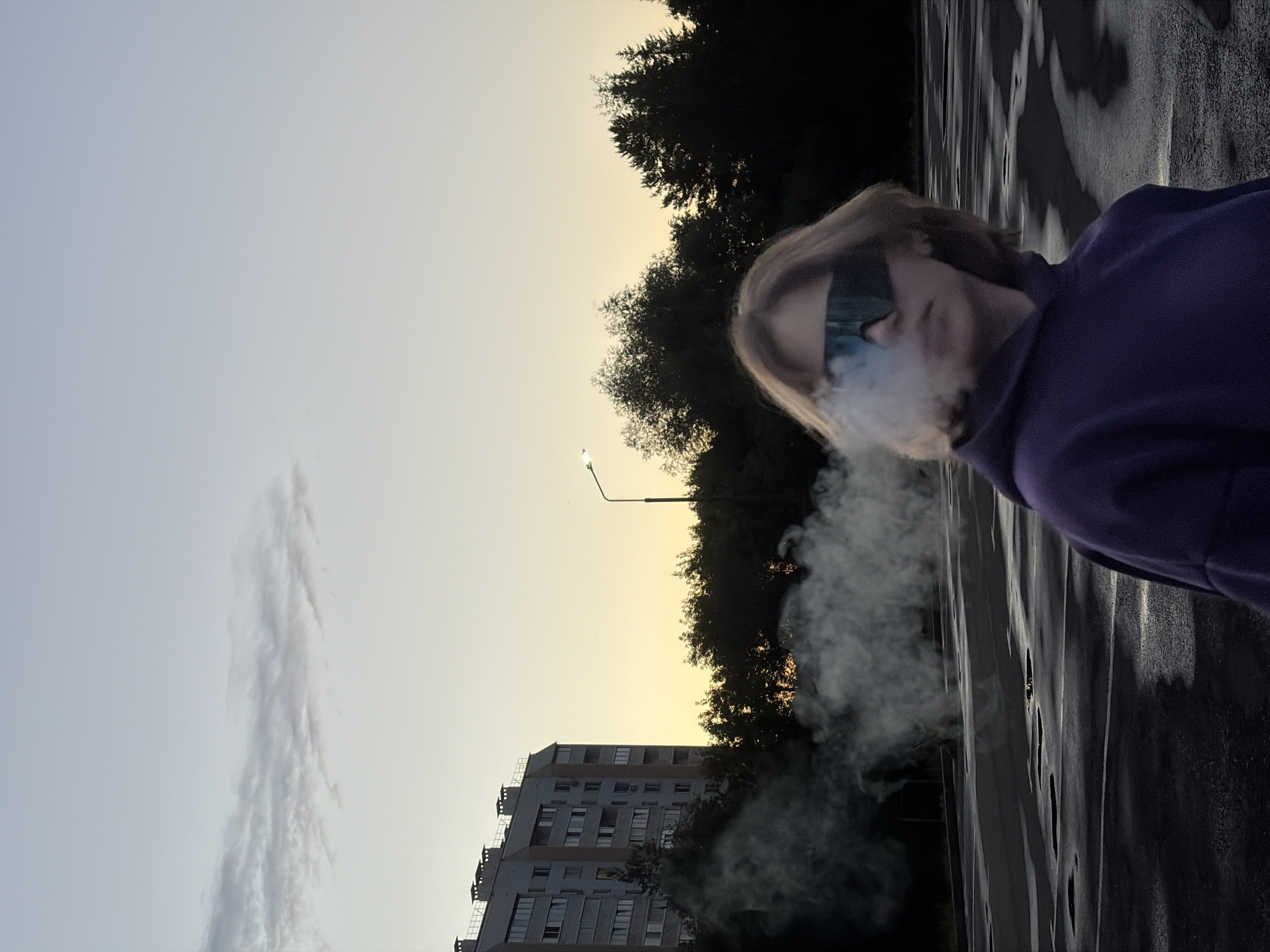 The image shows a person wearing a purple hoodie and reflective sunglasses, exhaling a cloud of smoke or vapor. The person is in the foreground, with their face partially obscured by the smoke. The background features a street lamp, trees, and a tall apartment building. The sky is a soft gradient of pale yellow and light blue, suggesting either sunrise or sunset. The ground appears to be wet and reflects the light, possibly indicating recent rain. The overall mood is somewhat mysterious, with the smoke adding an element of intrigue to the scene. The composition is vertical, with the person on the right side of the frame.
