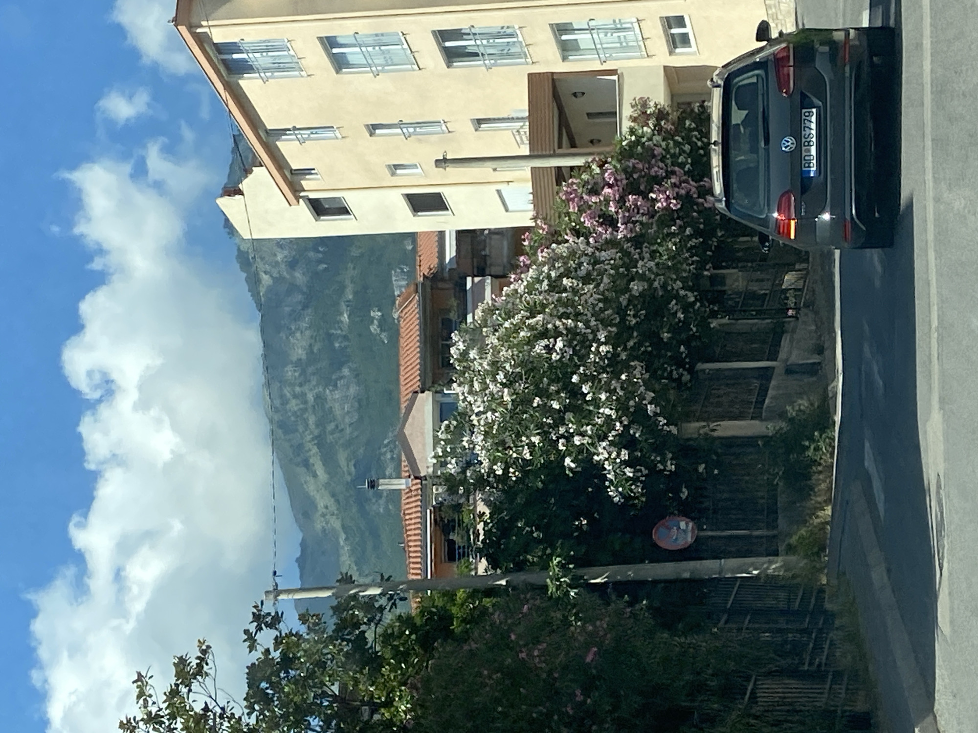 In Budva, Montenegro, a dark gray Volkswagen station wagon with license plate BD BS779 is parked on a paved road. The car is facing away from the camera. To the left of the road, a black metal fence runs along a dirt embankment. Behind the fence, lush green foliage and blooming trees with white and pink flowers are visible. A tall, thin utility pole stands near the fence. Further back, residential buildings with terracotta roofs and light yellow facades rise up. The buildings have multiple windows and balconies. In the background, a rugged, green mountain range stretches across the horizon under a bright blue sky dotted with white clouds. A circular traffic sign with red and blue elements is mounted on a pole near the fence. The overall scene depicts a street in a residential area, likely in Budva, given the license plate and architecture.