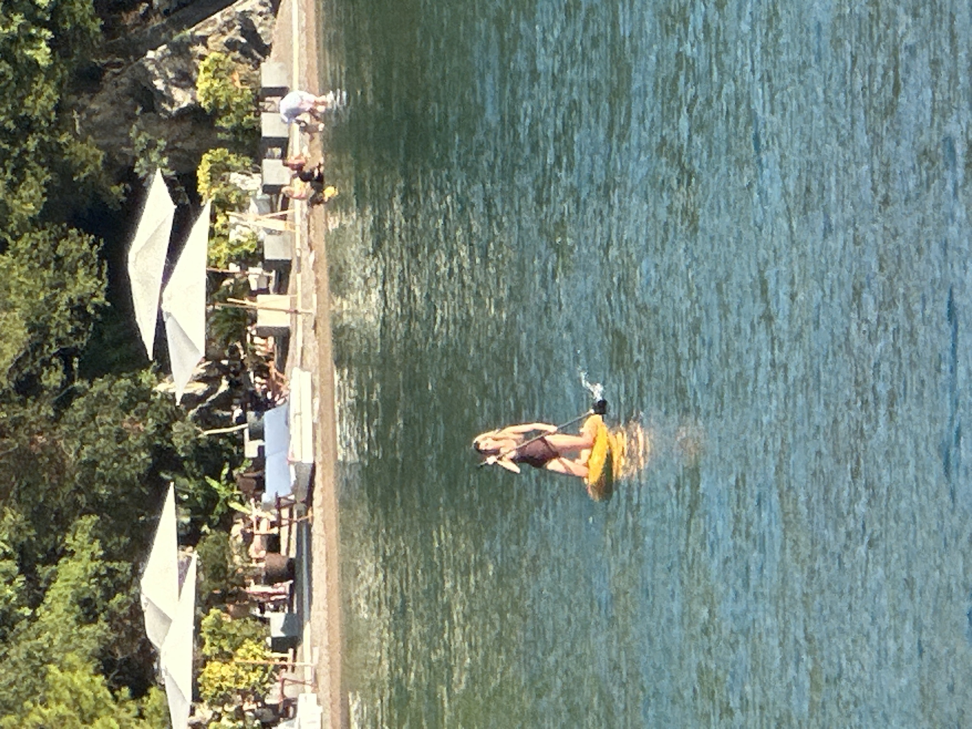 A woman is paddleboarding in the turquoise waters of the Adriatic Sea, near the coast of Budva, Montenegro. She is wearing a dark swimsuit and kneeling on a yellow paddleboard, holding a paddle. In the background, several people are relaxing on lounge chairs under white umbrellas on a sandy beach. Behind the beach, lush green trees and rocky cliffs form a natural backdrop. The sun is shining, casting reflections on the water.