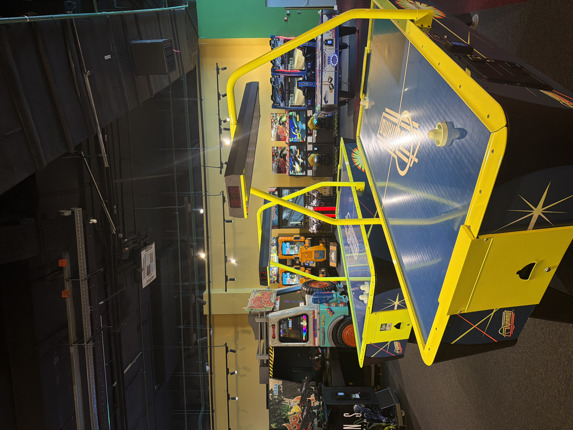 The image shows an indoor arcade. In the foreground, there are two air hockey tables with blue playing surfaces and yellow trim. The tables have digital score displays and mallets. In the background, various arcade games are visible, including racing games with driver cockpits and a Sega arcade machine. The walls are painted in bright colors, with yellow and green dominating. The ceiling has exposed pipes and lighting fixtures. The floor is carpeted. There are no people visible in the image.