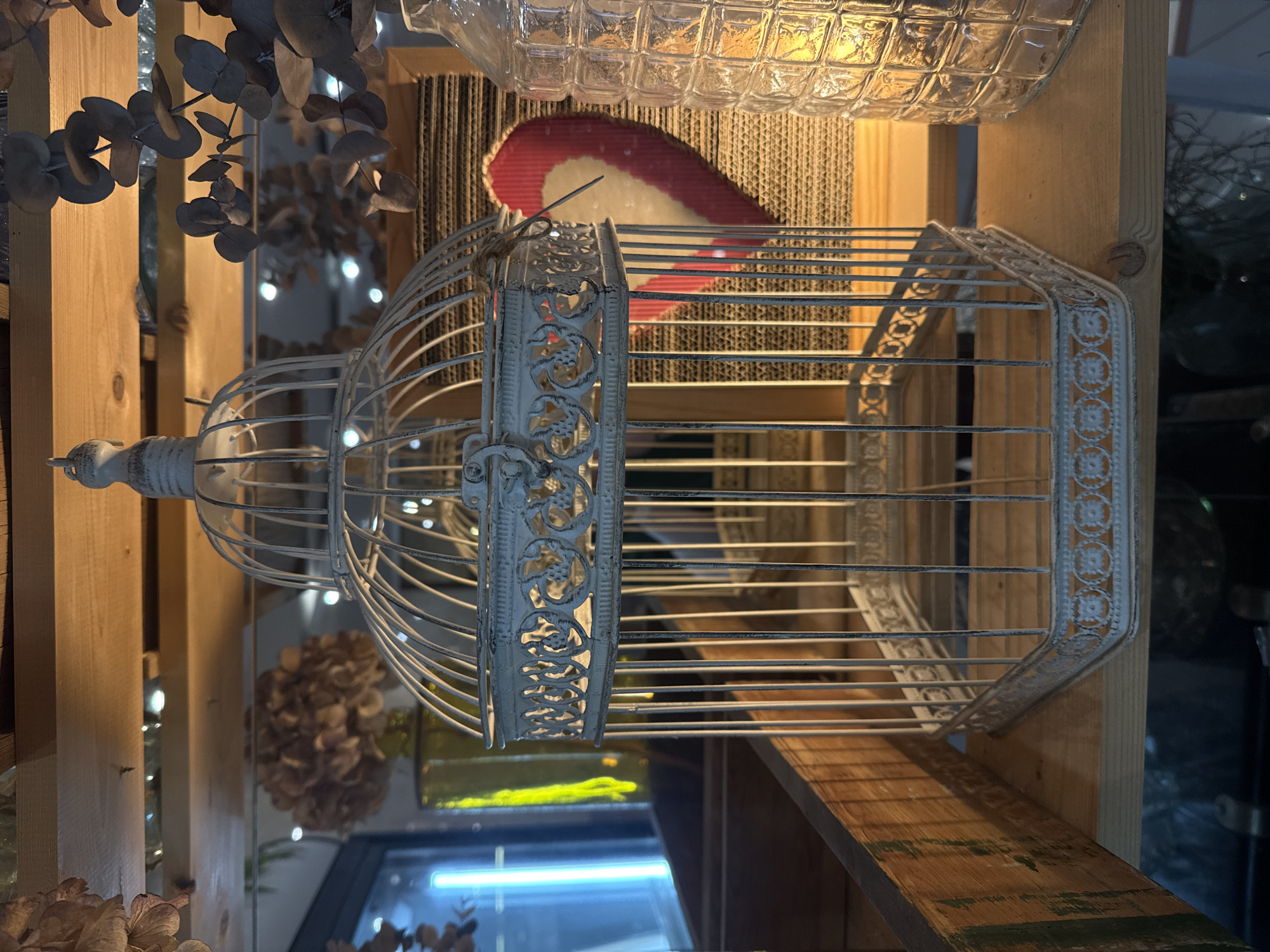 The image shows a white, vintage-style birdcage on a wooden shelf. The shelf appears to be part of a larger structure, possibly a display or shelving unit in a store. The background includes other decorative items such as dried flowers and a textured cardboard element, as well as a glass bottle. The location is not discernible from the image, and no information suggests the location is in Saint Petersburg, Russia. No people or actions are visible.