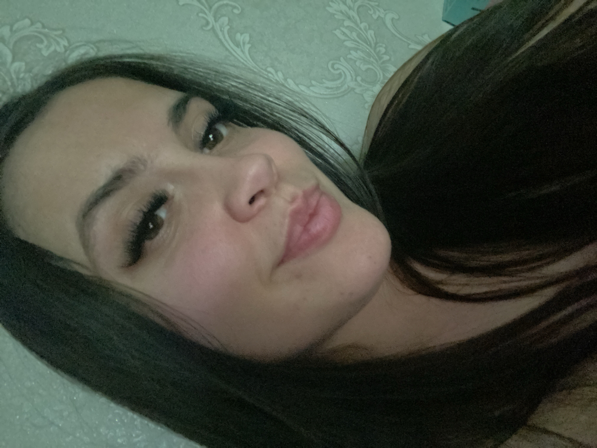 The image is a close-up selfie of a woman with long, dark brown hair. She is lying down, with her face tilted slightly towards the camera. She has brown eyes and full lips. She is wearing false eyelashes. The background is a light-colored wall with a patterned wallpaper. The location is not discernible from the image. There is no evidence to suggest the location is in the city of Chaikovsky, Russia.