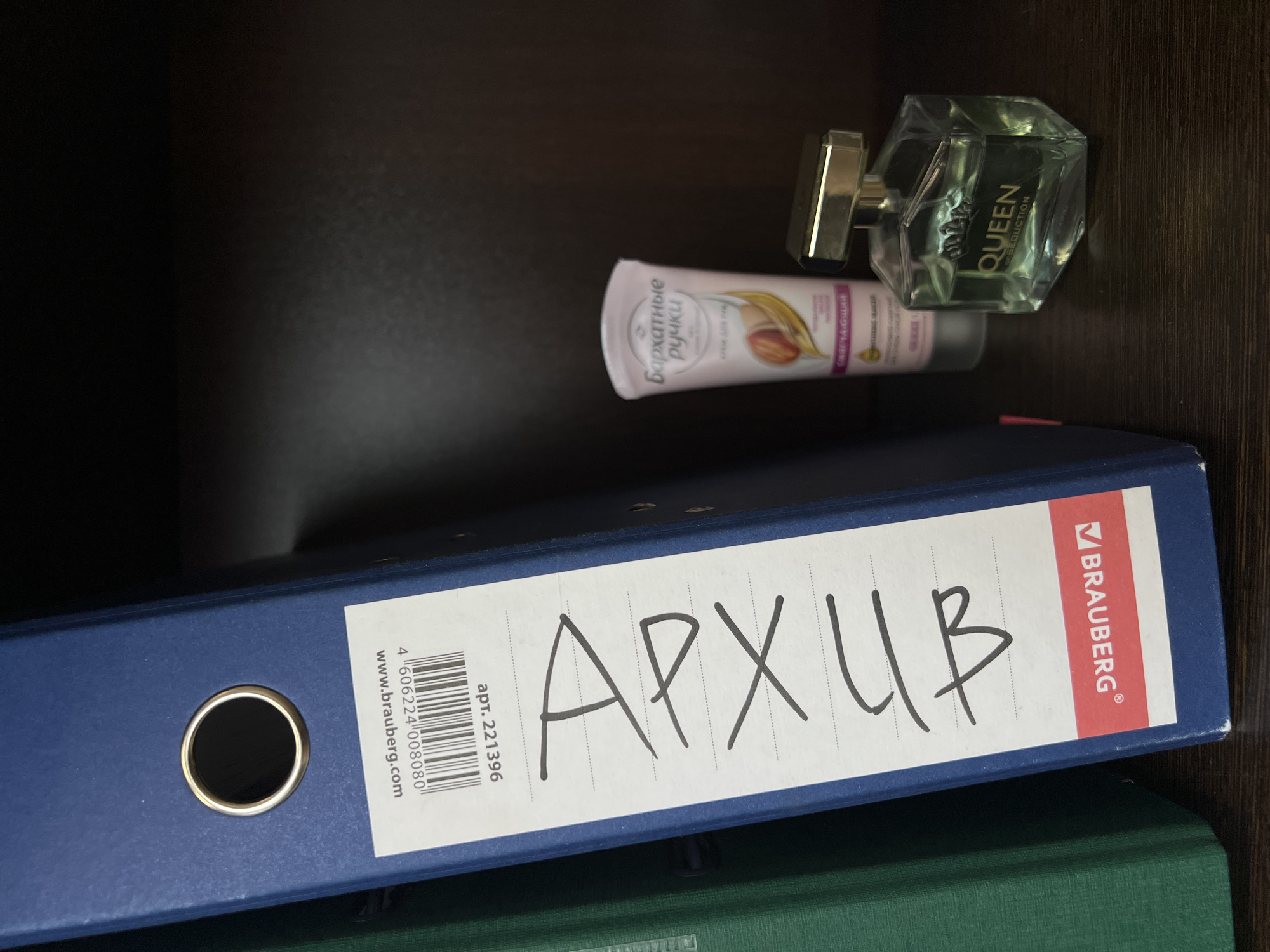 The image shows a shelf with several items. In the upper right, there is a bottle of perfume, labeled "QUEEN SEDUCTION." To the left of the perfume is a tube of hand cream with the Russian text "Бархатные ручки" (meaning "Velvet Hands"). In the foreground, there is a blue Brauberg brand binder with the Russian word "Архив" (meaning "Archive") written on it in black marker. The binder appears to be stacked on top of another object, partially visible at the bottom. It is impossible to determine the location in the city of Perm, Russia based on the visual information.