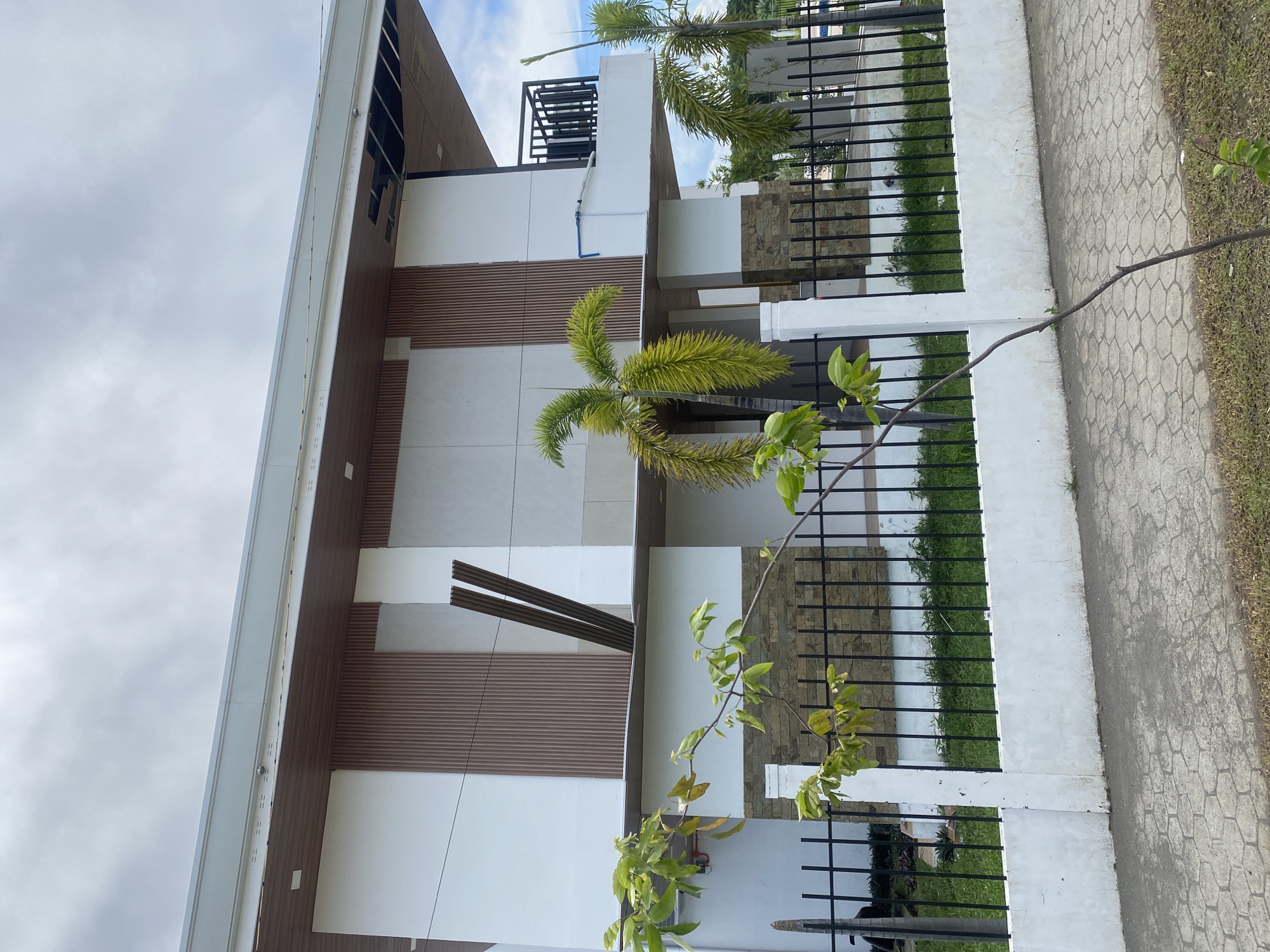 A partially constructed two-story modern house is seen from the street in Davao City, Philippines. The house features white walls with horizontal wooden paneling and some stone veneer. A black metal fence with a white concrete base separates the property from the sidewalk, which is paved with hexagonal concrete blocks. Several palm trees are planted in front of the house. The sky is overcast with clouds. The house appears to be incomplete, with exposed ceiling elements on the second floor and some construction materials leaning against the exterior wall.