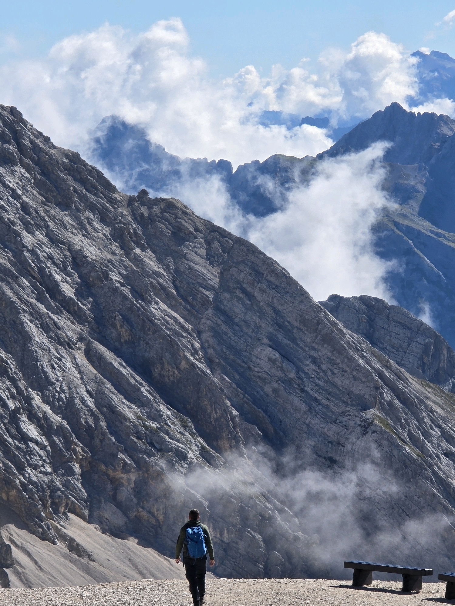 The image depicts a man with a blue backpack walking on a gravel path in a mountainous landscape. The man is facing away from the camera, walking towards a steep, rocky mountain face that dominates the left side of the frame. Wisps of clouds or mist are seen clinging to the mountain slopes and in the background, partially obscuring jagged peaks. The sky above is blue with scattered white clouds. In the foreground, to the right of the path, a wooden bench is visible. The scene appears to be in a high-altitude, natural environment. The location is not specified as being in the city of Garmisch-Partenkirchen, Germany, but the landscape is consistent with alpine regions.