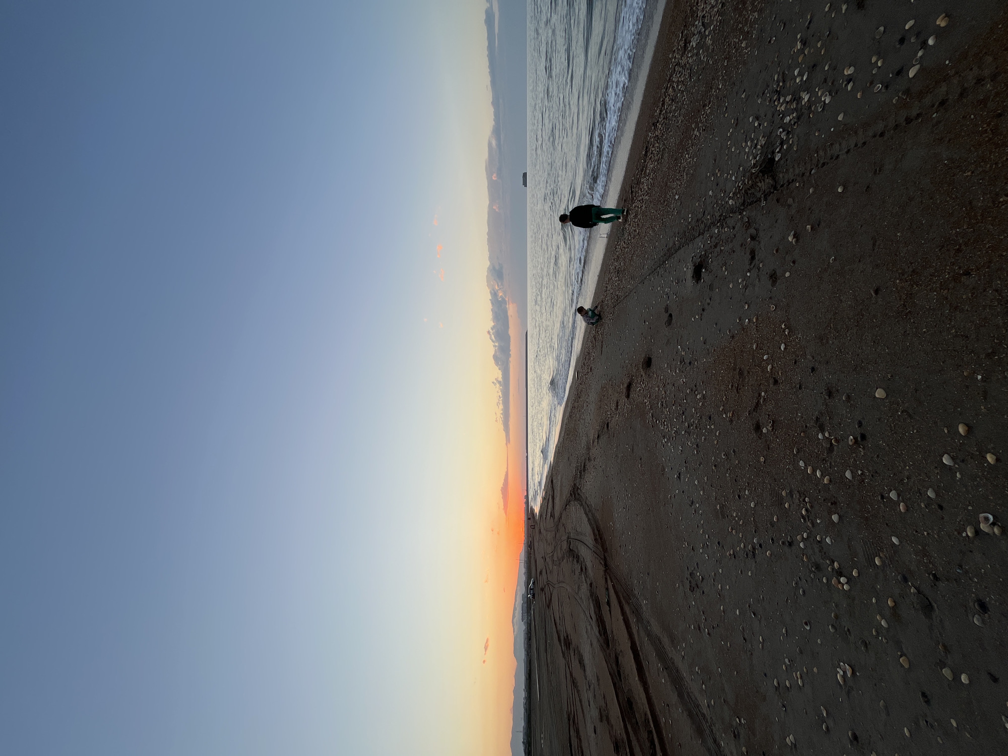 The image shows a beach in Kaspiysk, Russia, at sunset. The sky transitions from blue to orange near the horizon. Two individuals are present on the beach near the water's edge. One is standing and appears to be looking out at the sea, and the other is smaller and closer to the ground, possibly playing in the sand. Tire tracks are visible in the sand, suggesting vehicular activity on the beach. The beach is dark, with pebbles and shells scattered on the surface. The sea is calm, with gentle waves lapping at the shore. A ship is visible in the distance on the water.