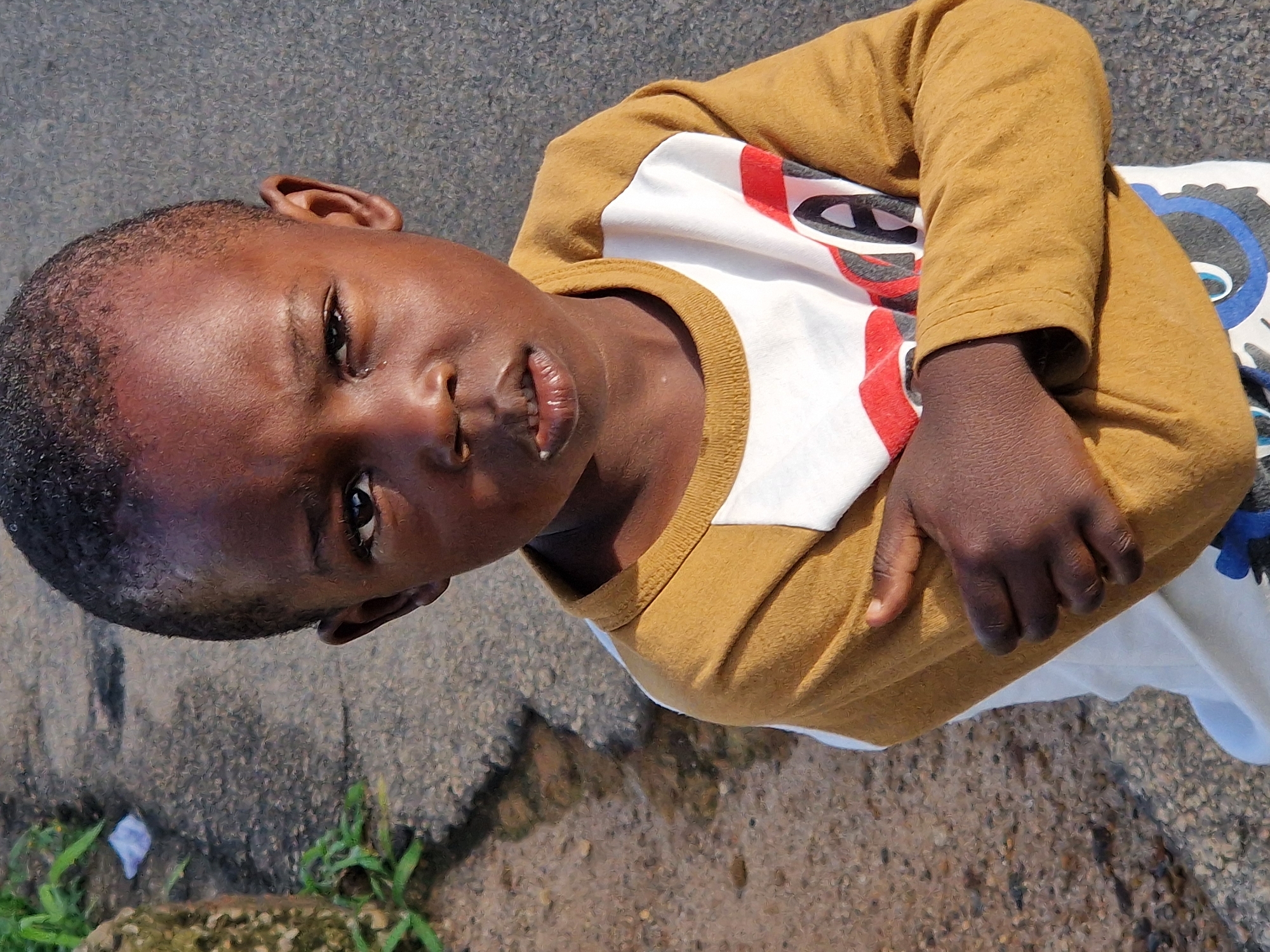 A medium shot, eye-level outdoor photograph shows a young Black boy with short hair standing on a paved surface. The boy is looking directly at the camera with a serious expression. He is wearing a long-sleeved mustard yellow shirt over a white shirt with a graphic print. His left arm is crossed over his chest, and his right arm is bent at the elbow. The background consists of a textured gray paved surface, with a small white object and some green foliage on the left side of the frame. The lighting suggests it is daytime.