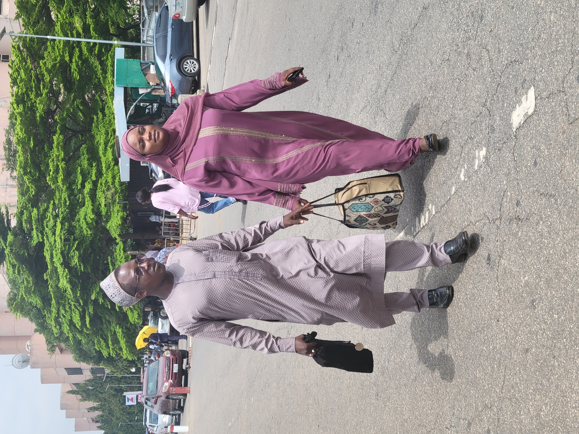 The image displays two individuals, a woman and a man, walking together on a paved road or parking lot in what appears to be a sunny urban environment, consistent with Abuja, Nigeria. The woman is on the left, dressed in a flowing purple outfit, likely an abaya or jilbab, adorned with gold or beige embroidery, and a matching head covering (hijab). She holds a mobile phone in her right hand and shares a patterned beige and blue shoulder bag with the man.
The man, positioned to her right, wears a light grey or beige patterned agbada-style tunic and trousers, along with a matching cap (kufi) and sunglasses. He holds a small black pouch or bag in his left hand, while his right hand also holds the patterned bag carried by the woman. Both are wearing dark shoes.
The background shows a busy street scene with several vehicles, including parked cars (a grey sedan, a red SUV) and a green tricycle (keke napep). Lush green trees line the street, and multi-story buildings are visible further back. Other people can be seen in the distance. The bright daylight casts clear shadows on the cracked asphalt surface, indicating midday or early afternoon. A notable detail is an "ATM" logo visible on a structure in the background. The overall scene depicts a casual walk or commute through a public area.