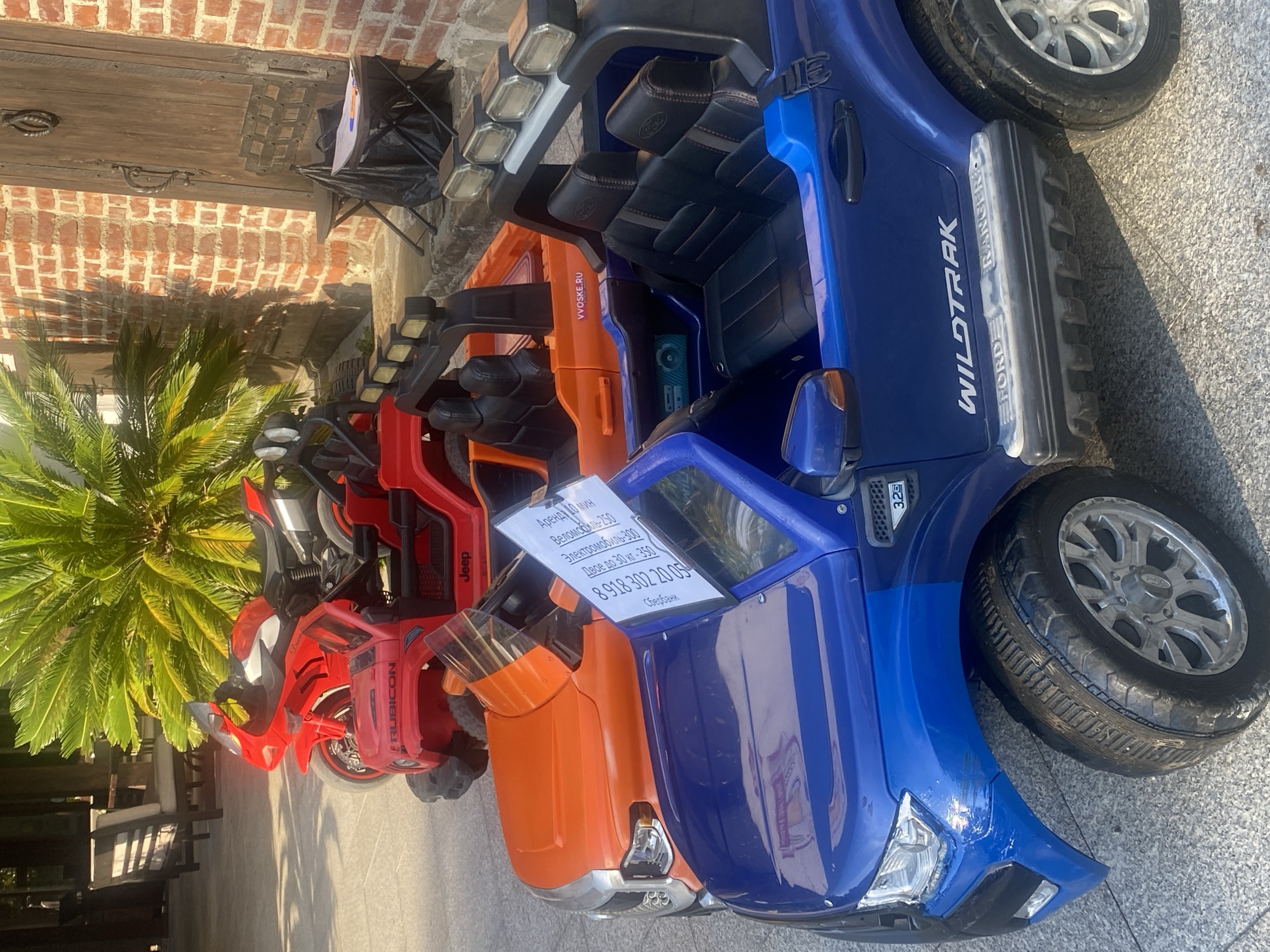 This media file shows three toy vehicles lined up outdoors. The vehicles appear to be electric ride-on cars for children, designed to resemble real vehicles. The leftmost vehicle is red with "RUBICON" and "Jeep" branding, suggesting it's a replica of a Jeep Rubicon. Next to it is an orange vehicle. The rightmost and largest vehicle is blue with "WILDTRAK" and "FORD RANGER" branding, indicating it's a replica of a Ford Ranger Wildtrak.
A white sign is attached to the orange vehicle, displaying text in Russian. The text includes rental information: "Аренда 10 мин" (Rental 10 min), "Веломобиль-250" (Velomobile-250), "Электромобиль-300" (Electric car-300), and "Двое до 30 кг-350" (Two up to 30 kg-350). A phone number, "8 918 302 20 05", and "Сбербанк" (Sberbank) are also visible on the sign. Another website address, "VVOSKE.RU", is present on the blue vehicle.
The vehicles are parked on a paved surface, likely outdoors, with a brick wall and a wooden door in the background. To the left, a portion of a palm tree is visible, suggesting a warm climate. The lighting indicates it is daytime, with shadows cast on the ground. The scene appears to be a rental stand or an outdoor play area. No people are visible in the image, but the presence of children's ride-on toys implies they are the intended users. The overall impression is a commercial setup offering rental of these toy vehicles.
