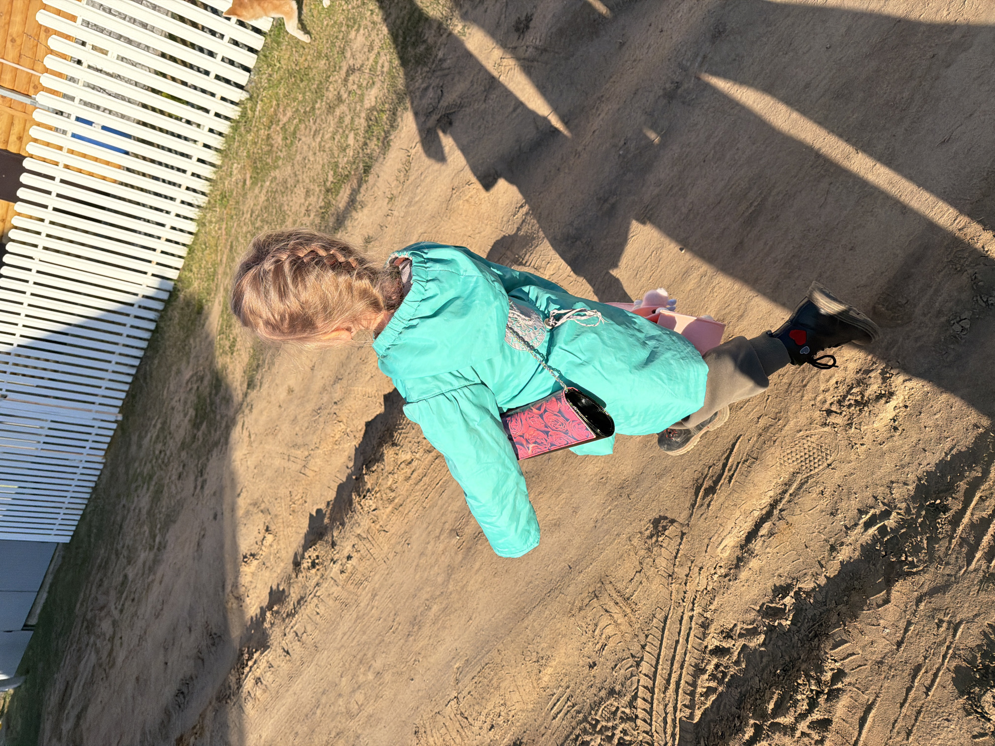 A young child is walking along a sandy path next to a white picket fence. The child is wearing a turquoise jacket, brown pants, and black boots with red heart accents. A small purse with a floral pattern hangs from the child's shoulder. The ground is sandy with visible tire tracks. The image shows a sunny day. I don't have additional information about this image that would tell me where specifically in the city of Chaikovsky, Russia that the image was taken.