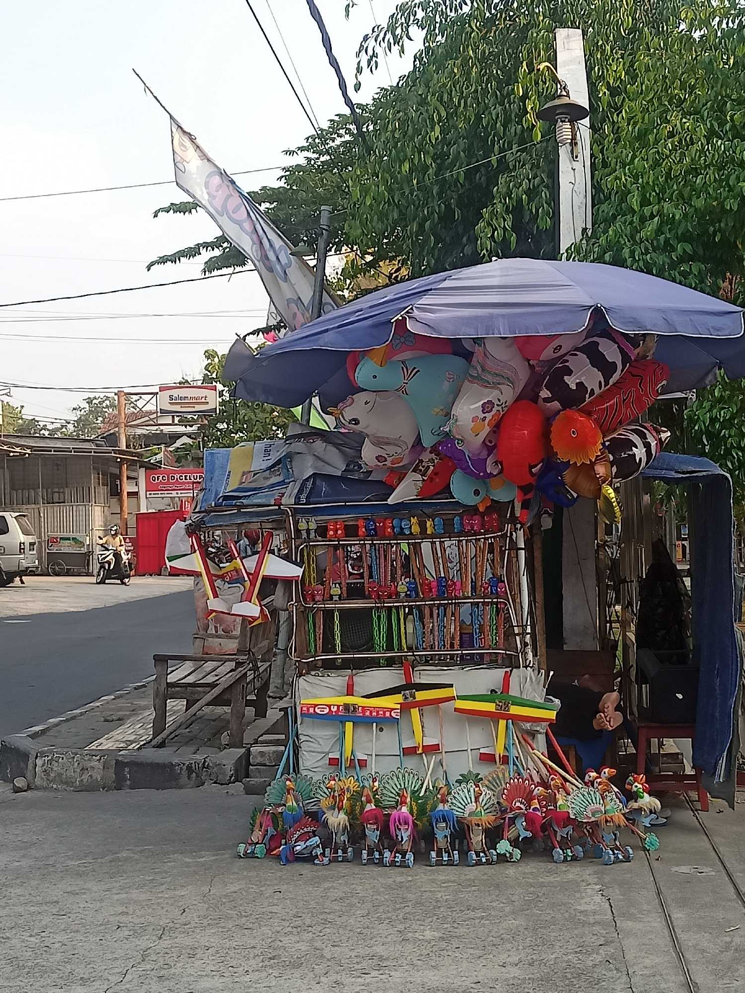 This is an eye-level, outdoor, daytime, medium shot of a street-side stall in Girimargo, Indonesia. The stall is piled high with colorful toys, including balloons and small figurines. A blue umbrella shades the top of the stall. Behind the stall, lush green trees line the street, and a utility pole with a light fixture is visible. The street is paved, and a sidewalk with a wooden bench is present. In the background, buildings and a sign for "Salemamart" are visible. A person on a motorcycle drives down the street. The overall scene depicts a typical street market in a town.