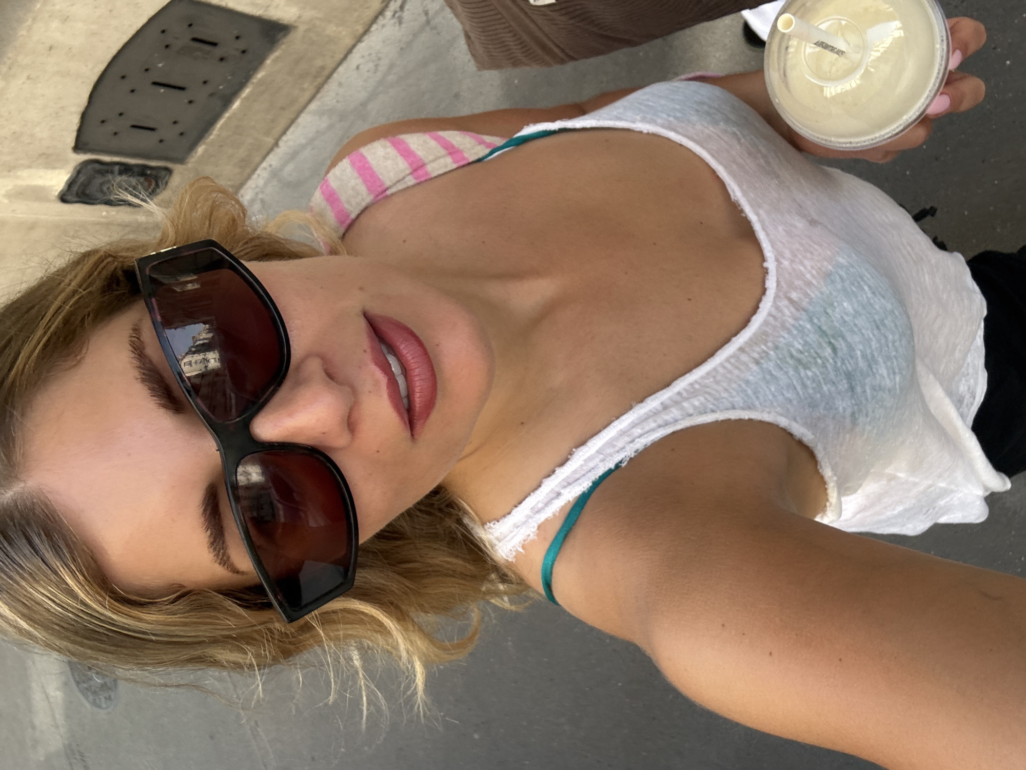 A woman takes a selfie. She is wearing sunglasses, a white tank top, and a striped bra strap is visible. She is holding a drink in a plastic cup with a straw. In the lens of her sunglasses is the reflection of a building. The photo was taken on a street with concrete, and a grate in the concrete can be seen in the background. The reflection in the woman's sunglasses shows a building. Based on the architecture visible in the reflection, the building is most likely located in Paris, France.