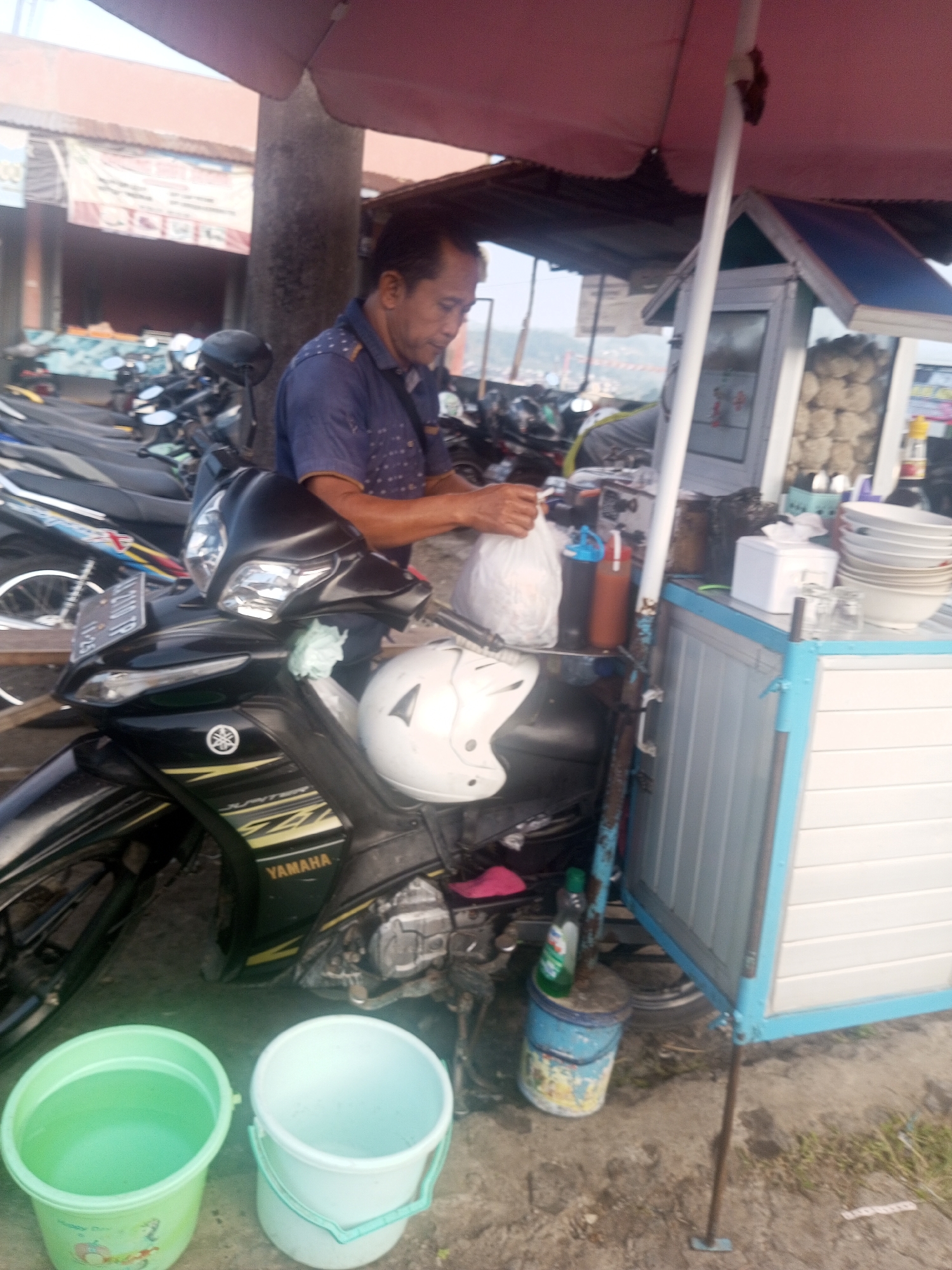 A middle-aged Indonesian man, wearing a blue patterned short-sleeved shirt, stands next to a black Yamaha motorcycle, holding a white plastic bag in his right hand. He is positioned at an outdoor food stall, which has a blue and white striped facade. The stall displays stacks of white bowls and cups on its counter. Behind the man and the motorcycle, other motorcycles are parked in a row. In the foreground, two plastic buckets, one light green and the other white, are placed on the ground. The scene appears to be a street-side vendor operation, likely in the city of Kalibeber, Indonesia. The ground is paved with concrete and dirt.