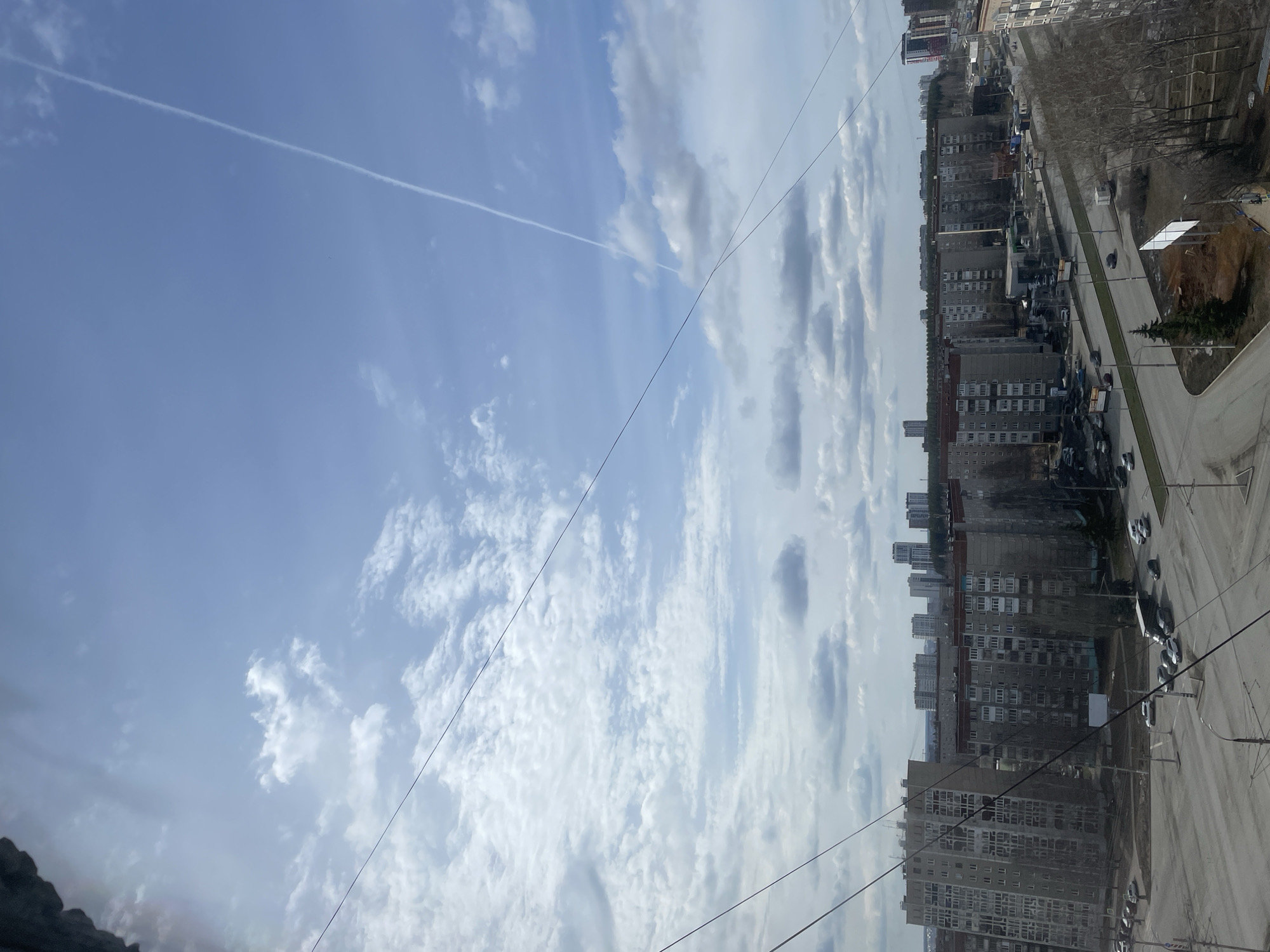 The image shows an urban scene in Izhevsk, Russia, with a focus on a view from an elevated position, possibly from a high-rise building or a window. The sky occupies a significant portion of the frame, featuring a blue expanse interspersed with white clouds and a contrail. Below the sky, a cityscape unfolds, composed primarily of multi-story residential buildings. These buildings are predominantly gray and utilitarian in appearance, reflecting typical Soviet-era architecture. A street runs alongside the buildings, showing parked cars and sparse vegetation. The scene is captured from an unusual angle as the image is slightly rotated, but the composition emphasizes the contrast between the sky and the urban landscape. Electrical wires cut across the frame in the foreground. There are no visible people in the image. The overall environment suggests a moderately populated area within Izhevsk.