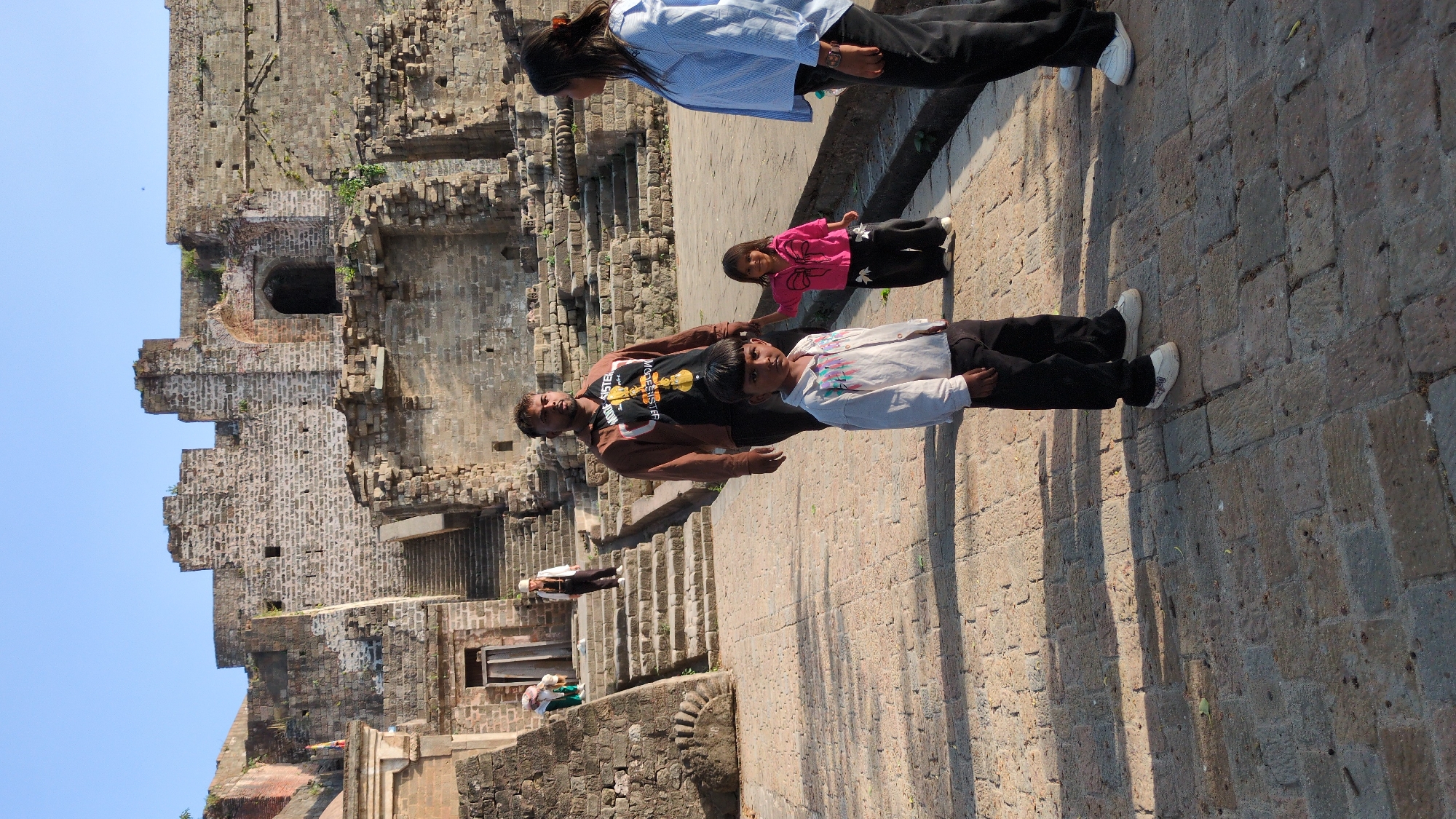 The image captures a scene at the ruins of the Qila Mubarak fort in Kaswara, India. Four individuals are visible: a woman in a blue shirt and dark pants at the top, a young girl in pink, and a man in a tank top with a young boy in a white shirt standing beside him. They are all standing on a paved area, which casts long shadows. The ruins of the fort consist of weathered stone walls and structures, with an open doorway and multiple levels, set against a clear blue sky. Steps lead up into the fort structure. Other people can be seen in the background.