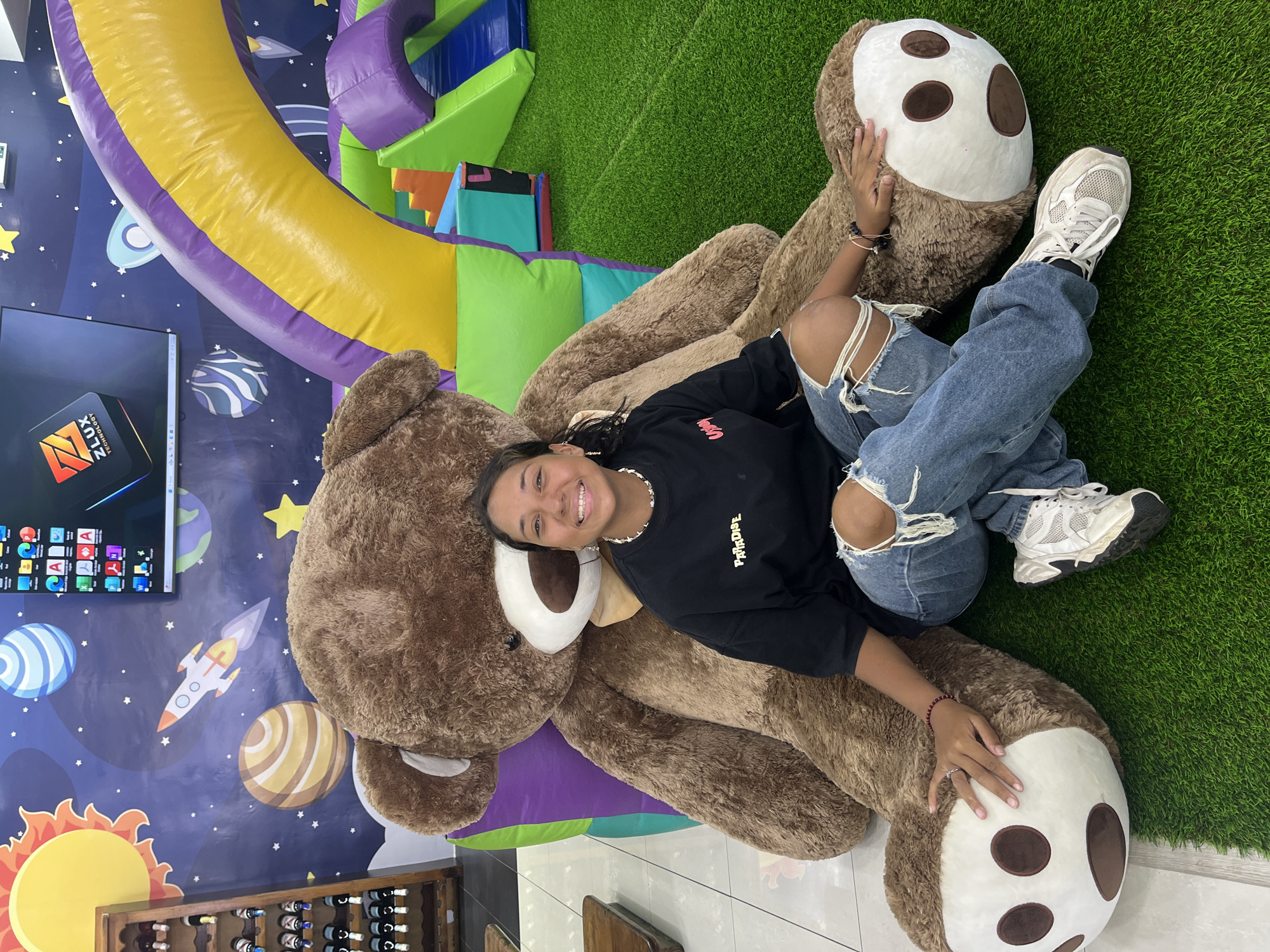 A young woman with dark hair lies on a large, brown teddy bear with a white and brown sloth face. She wears a black t-shirt with "Paradise" written on it, ripped blue jeans, and white sneakers. The location appears to be an indoor play area featuring a colorful, inflatable structure and a wall with space-themed graphics. To the left, there is a television screen and a wine rack. The flooring transitions from a faux-grass green to a light-colored tile. The city and country are not evident in the picture.