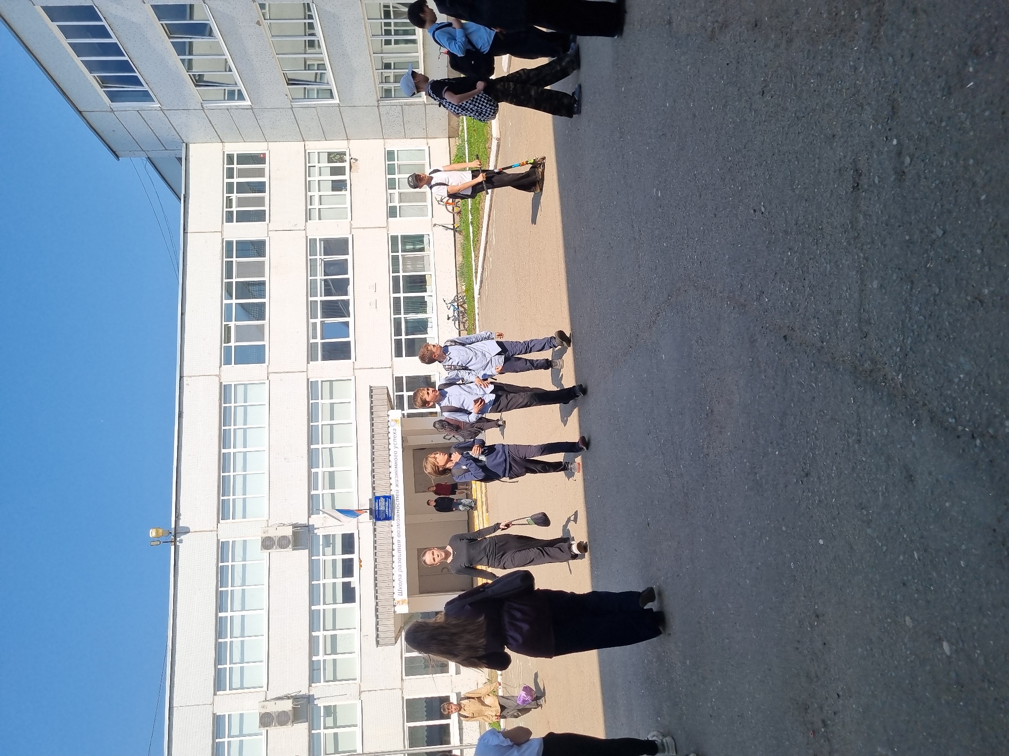 The image shows a group of people standing outside a multi-story building in Chaikovsky, Russia. The building is light-colored with numerous windows and several air conditioning units. A street light is mounted on the building. The sky is clear and blue. The ground is paved with asphalt. The people are of various ages and are dressed in casual clothing. Some are wearing school uniforms. One person is holding a bouquet of flowers. Others appear to be waiting or standing in line. A person is holding a scooter.
