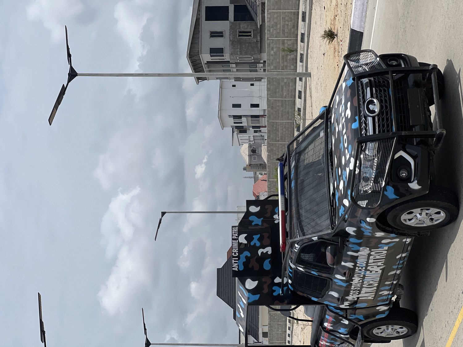 The image shows a Lagos State Police Command Anti-Crime Patrol vehicle parked on an asphalt surface. The vehicle is black with a camouflage pattern of blue, white, and gray. "ANTI CRIME PATROL" is written vertically on the side of the vehicle, along with "LAGOS STATE POLICE COMMAND." The vehicle has a metal guard on the front and a metal cage-like structure behind the cab.
In the background, there are multi-story buildings under construction, mostly gray with visible brickwork. Several modern streetlights line the road. The sky is overcast with gray clouds. The scene appears to be located in a newly developed area within a residential zone. The location is in the city of Alaguntan, Nigeria.