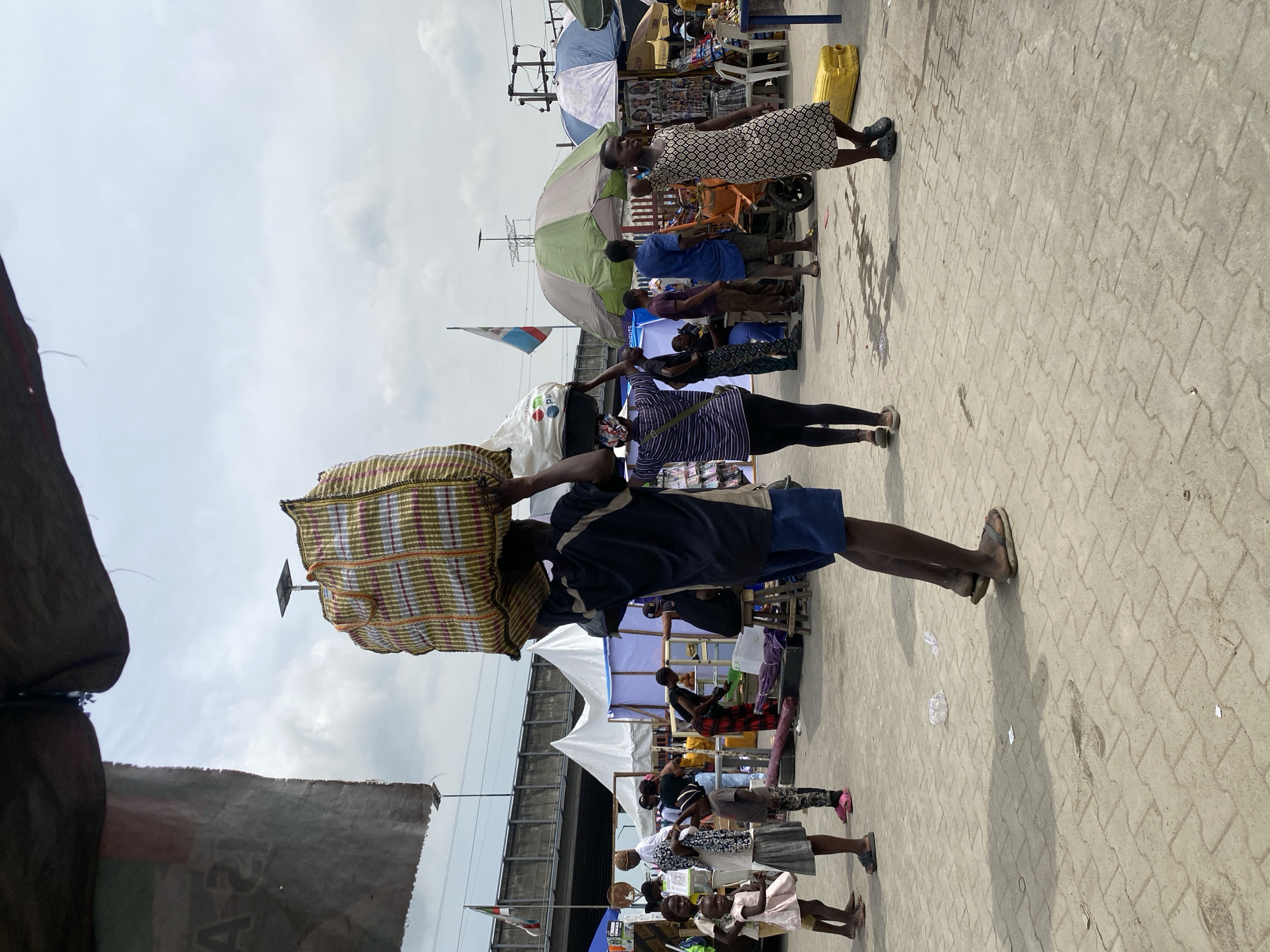 This image depicts a bustling outdoor market scene in Lekki, Nigeria, under an overcast sky. The ground is paved with interlocking concrete blocks, suggesting a designated public space or street.
In the foreground, a person walks towards the right, balancing a large, striped woven bag on their head. They wear a dark blue short-sleeved top, blue shorts, and sandals. To their right, another individual in a striped long-sleeved top and dark leggings appears to be interacting with a stall.
The midground is filled with various market activities and structures. Several people, including adults and children, are present. One woman in a white and black patterned dress stands near a stall shaded by a light green umbrella. Other market stalls, some covered by white canopies, display various goods, though the items themselves are not clearly discernible. A yellow plastic jerrycan rests on the ground near one of the stalls. Further back, an elevated structure, possibly a bridge or overpass, spans horizontally across the image. Utility poles and power lines are visible against the cloudy sky. A flag, likely the Nigerian flag (green and white), is visible on a pole to the left of the elevated structure.
The sky is a light grey, indicating an overcast or cloudy day, but with sufficient daylight for outdoor activity. Shadows are soft and not sharply defined. On a white bag hanging near the left edge, the text "A21" is partially visible. The overall scene conveys a busy, functional urban environment characteristic of a local market.