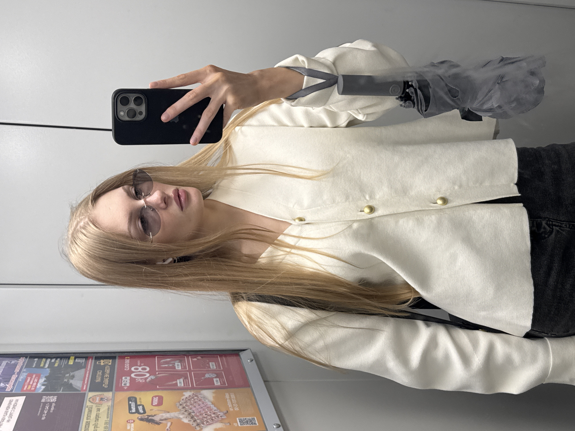 A young blonde woman with light skin and a fair complexion, wearing a white button-up jacket and dark jeans, is taking a mirror selfie in an elevator. She has on oval-shaped sunglasses and small white earbuds. In her right hand, she holds a black smartphone, capturing her reflection. She is also holding a closed grey umbrella by its handle, which is looped around her wrist. The elevator walls are a neutral grey color, and a framed advertisement is visible on the left wall. The overall environment suggests an urban setting, likely a residential building or a commercial establishment with elevator access. While the image does not provide specific indicators of the city of Perm, Russia, its general urban context is evident.