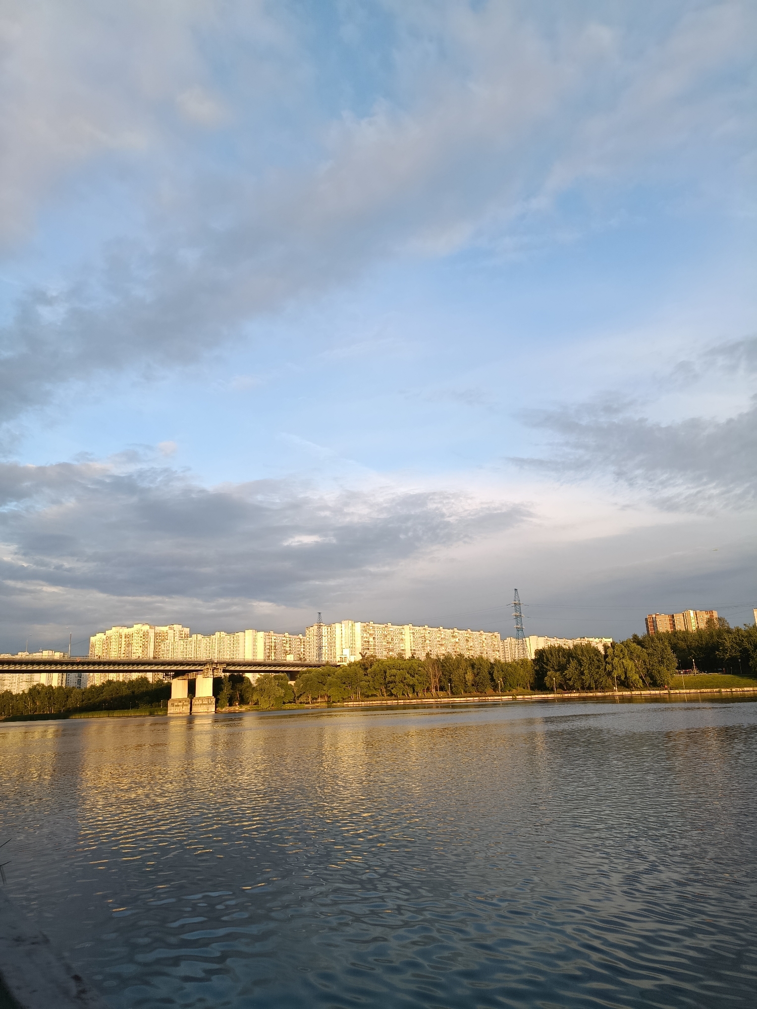 The image captures a wide shot of a river in Moscow, Russia, with a cityscape visible in the background. The sky is partly cloudy, with a mix of blue and grey hues, suggesting it might be late afternoon or early evening. On the far bank of the river, a long stretch of residential buildings with many windows dominates the skyline. A bridge, supported by concrete pillars, crosses the river in front of the buildings. Dense green trees line the riverbank, creating a natural buffer between the water and the urban structures. The surface of the river is textured with ripples, and the golden light of the setting sun reflects off the water, casting a warm glow on the scene. A few utility poles are also visible among the buildings. The overall impression is one of a calm, serene evening in a developed urban area.