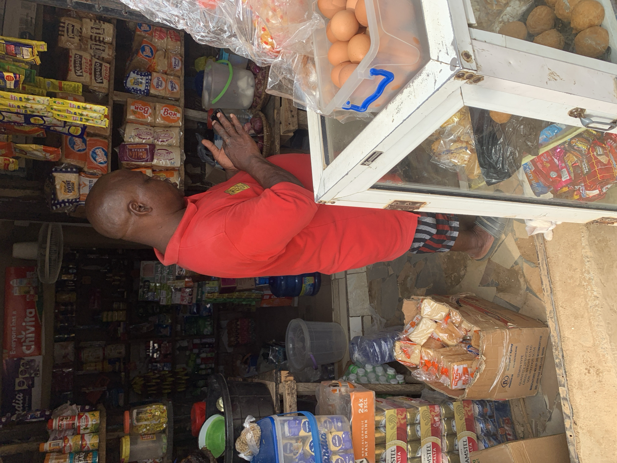 An overhead shot captures a man in a red polo shirt and striped shorts standing at a makeshift market stall in Abuja, Nigeria. He is holding a mobile phone in his hands, appearing to be engaged with it. The stall is laden with various goods, including packaged bread, canned beverages, and a clear plastic container filled with eggs. Behind him, shelves overflow with more products, and a ceiling fan is visible. To the right, a white display case shows more food items. The ground is a mosaic of brown and grey tiles.