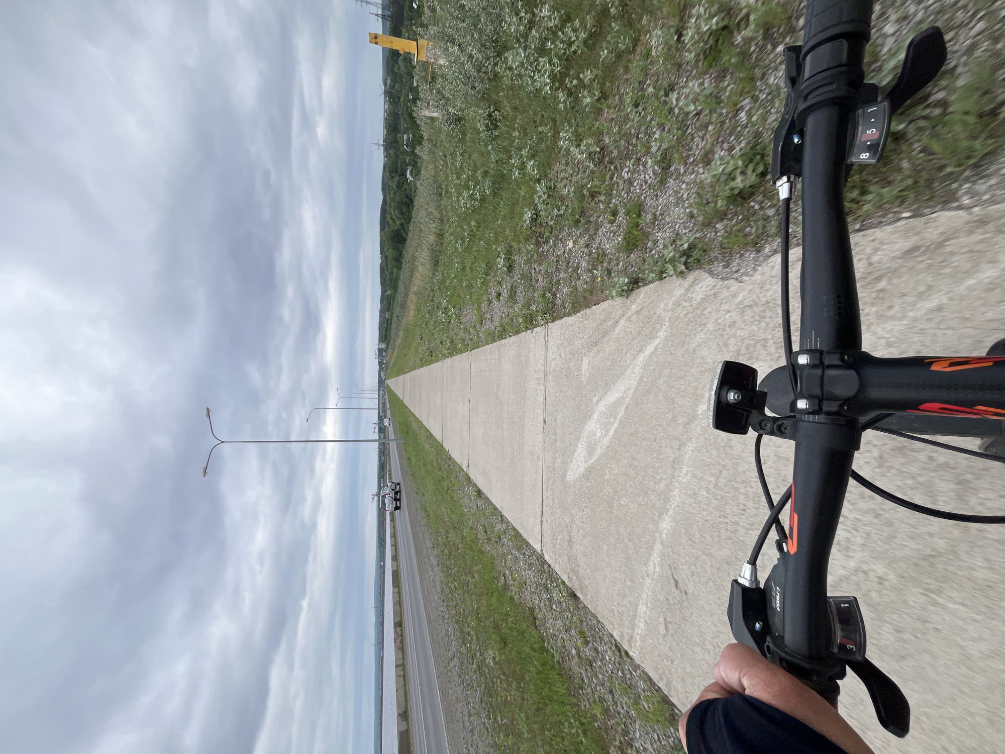 The image depicts a first-person perspective of someone riding a bicycle along a paved pathway. The pathway runs parallel to a road with some vehicle traffic, and beyond that, a body of water is visible. The environment is characterized by a cloudy sky, sparse vegetation along the roadside, and some distant buildings and industrial structures. Specifically, the location is identified as Chaikovsky, Russia, along the Kama River, a prominent geographical feature of the city. The cyclist's hand is visible on the handlebars, indicating the action of riding. The bicycle is equipped with a front light. The overall scene suggests an outdoor activity in an urban or semi-urban setting under overcast weather conditions.