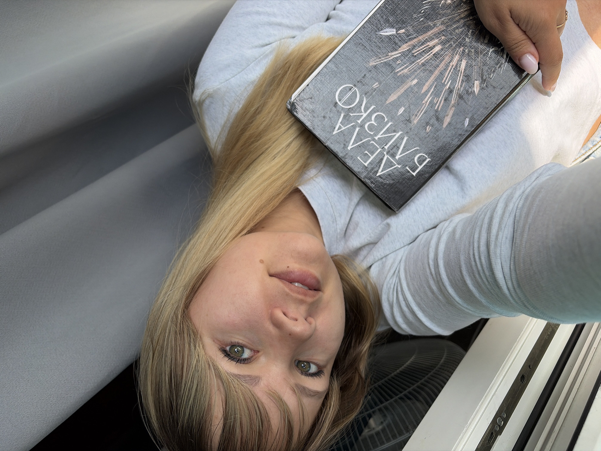 A selfie captures a young woman looking directly at the camera with a relaxed, slight smile in an indoor setting in Chaikovsky, Russia. She has long, straight blonde hair with bangs framing her face and notable light green or hazel eyes. A small mole is visible above her upper lip. She wears a light grey, long-sleeved top.
With her right hand, which has light pink or nude manicured nails and a simple ring on her ring finger, she holds a dark-covered book. The book's cover features a design resembling radiating light or an explosion, along with Cyrillic text clearly displaying "АЛЛА БАНКО" (ALLA BANKO).
The background to her right consists of a light grey, textured surface, possibly a curtain or wall. To her left, a dark, circular electric fan is partially visible, along with a white window frame or similar architectural element with dark accents. The scene is well-lit, indicating it is daytime. The woman's expression conveys a calm and casual demeanor.