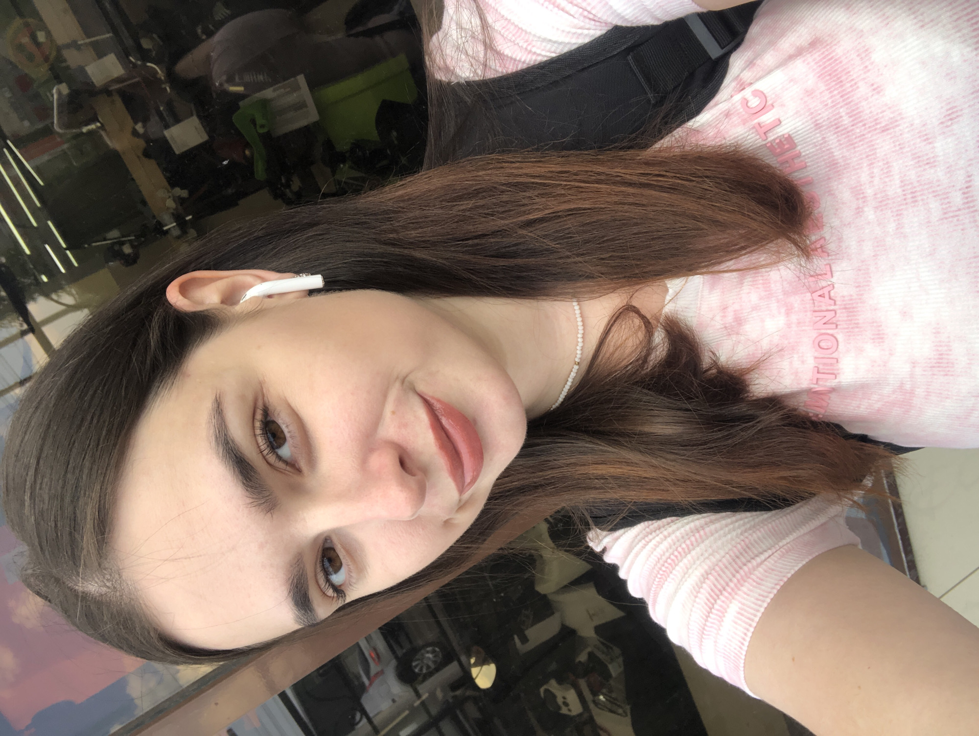 The image is a selfie of a young woman with long, brown hair and light skin. She is wearing a pink and white striped shirt and a black backpack. She has on white earbuds and a pearl necklace. She is looking at the camera and smiling. The background appears to be a reflection in a window or glass door, showing what looks like a store interior with shelves and displays. The woman is standing outdoors. It is not possible to determine her location in the city of Chaikovsky, Russia from the image alone.