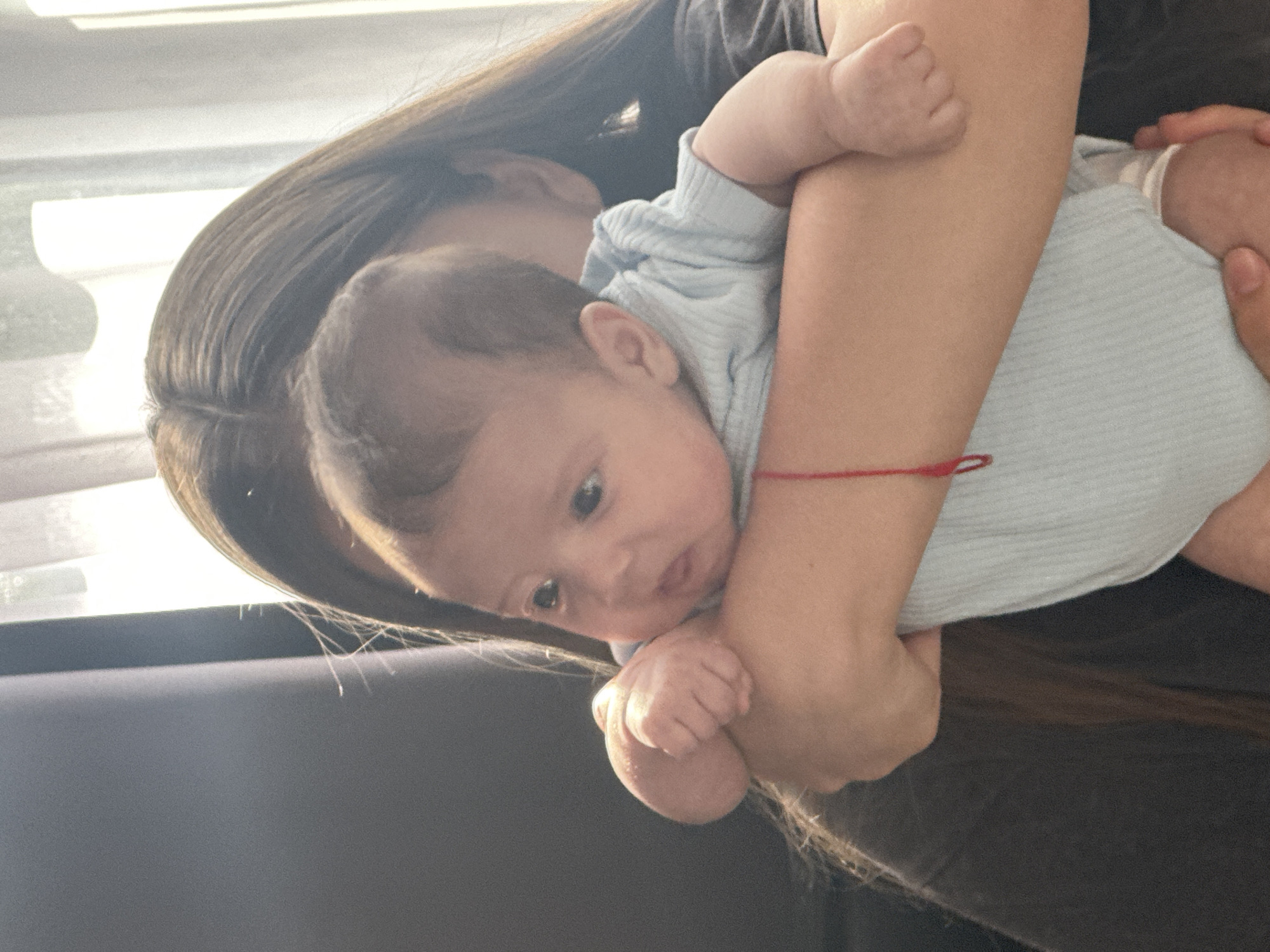 In Sochi, Russia, a person is holding a baby. The person, whose face is not visible, has long, dark hair and is wearing a dark t-shirt. The baby, who is facing to the left, is wearing a light blue onesie and has a small red string bracelet on their left wrist. They are being held close to the person's chest, supported by their arms. The background consists of a blurry gray surface on the left and sheer white curtains with sunlight streaming through on the right. The scene appears to be indoors, possibly in a home.