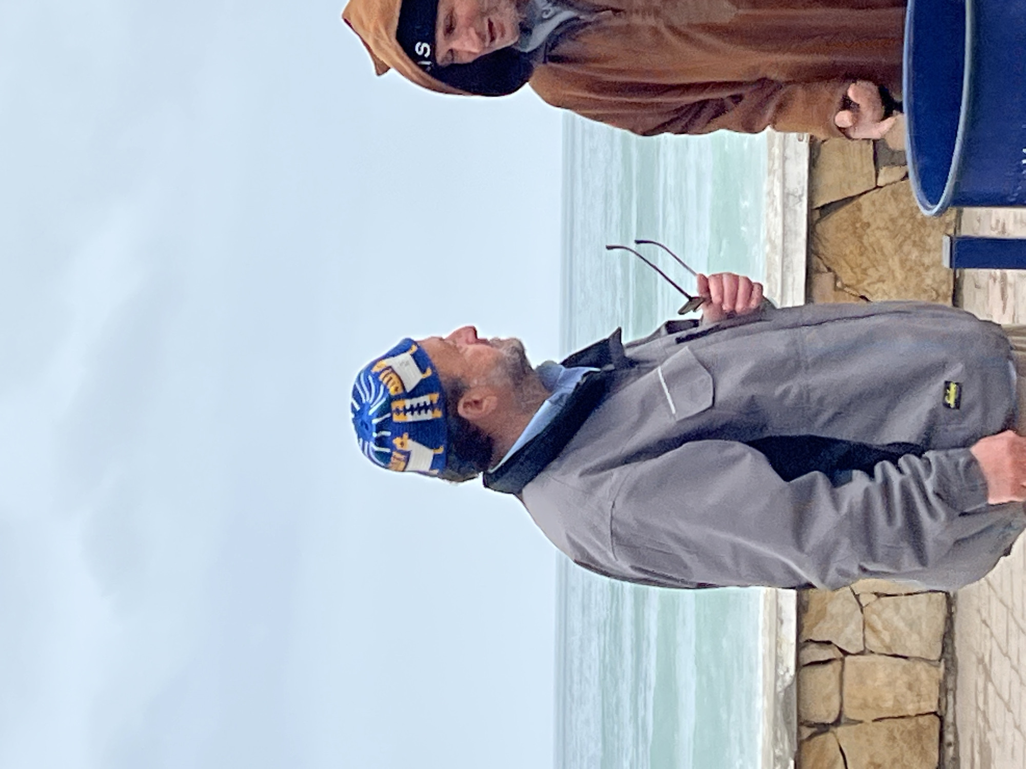 Two men stand on a paved promenade beside a large body of water under an overcast sky. The image is oriented sideways, but depicts an upright scene.
In the foreground, a man with a beard wears a blue, yellow, and white patterned beanie, a light blue collared shirt, and a grey jacket. A small yellow rectangular logo is visible on his right sleeve. He holds brown-framed sunglasses in his right hand and looks upwards with a slight smile.
Behind him and slightly to the right, a second man wears a brown hooded jacket and a black beanie with a distinct white "S" logo. He has facial hair and appears to be looking towards the first man. His hands are partially visible, one near a blue bin or container that is cropped on the far right.
The setting is a tranquil coastal area, consistent with a promenade along the sea in M'Diq, Morocco. A calm, light greenish-blue body of water stretches across the background, separated from the paved area by a low stone wall or barrier made of light-colored blocks. The sky is a uniform, light grey, indicating an overcast day.
The men appear to be engaged in conversation or observing their surroundings. The cool weather is suggested by their attire, including jackets and beanies. The pattern on the foreground man's beanie includes stylized animal heads and geometric motifs.