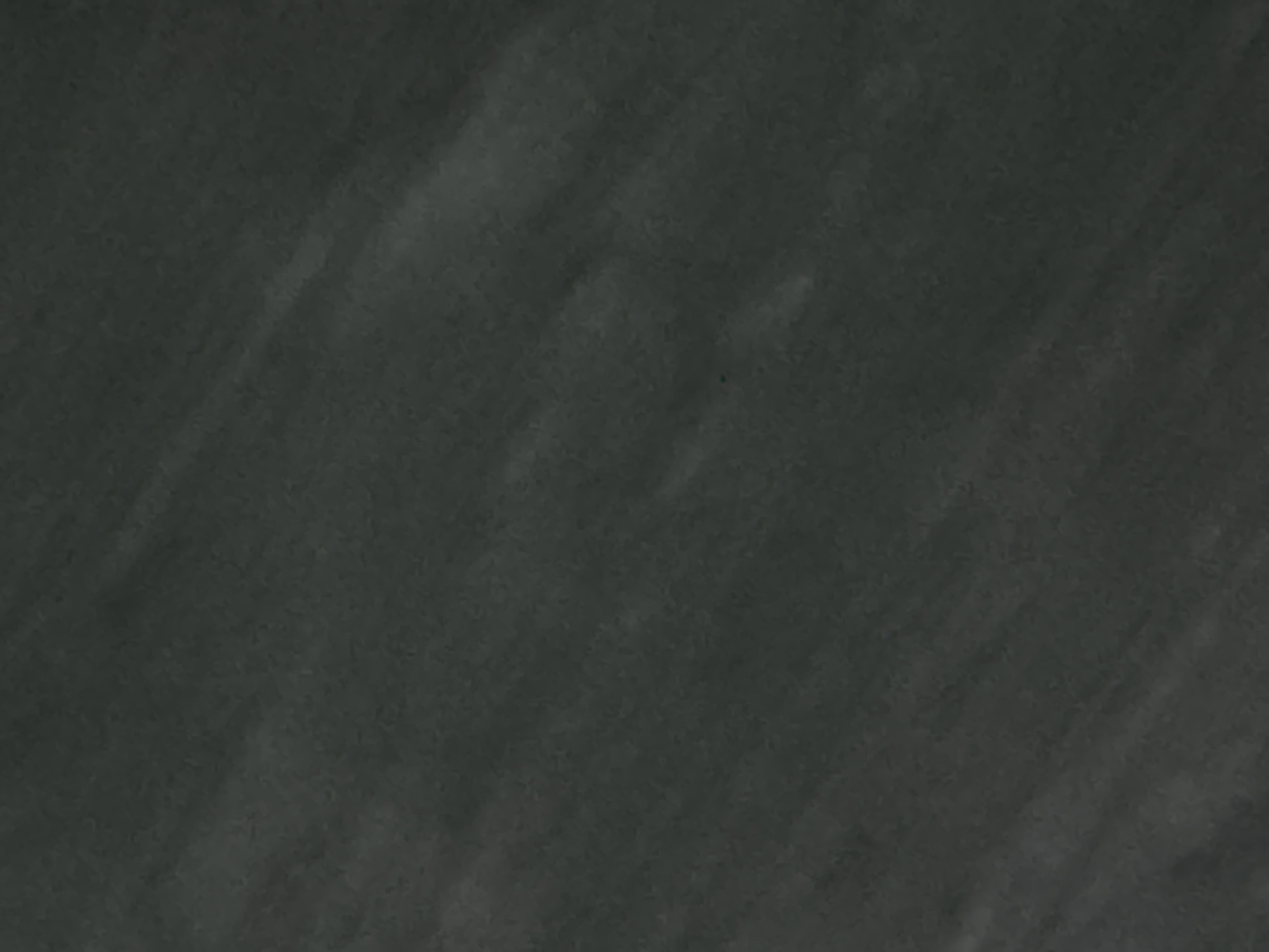 The image is a close-up, abstract shot of a dark, textured surface. The surface appears to be a wall or a fabric, with visible brush strokes or a rough grain. The lighting creates subtle variations in shade, suggesting a directional light source. There are no discernible objects, people, or any clear indication of a specific location within Navbahor, Uzbekistan. The overall impression is one of muted tones and abstract texture.