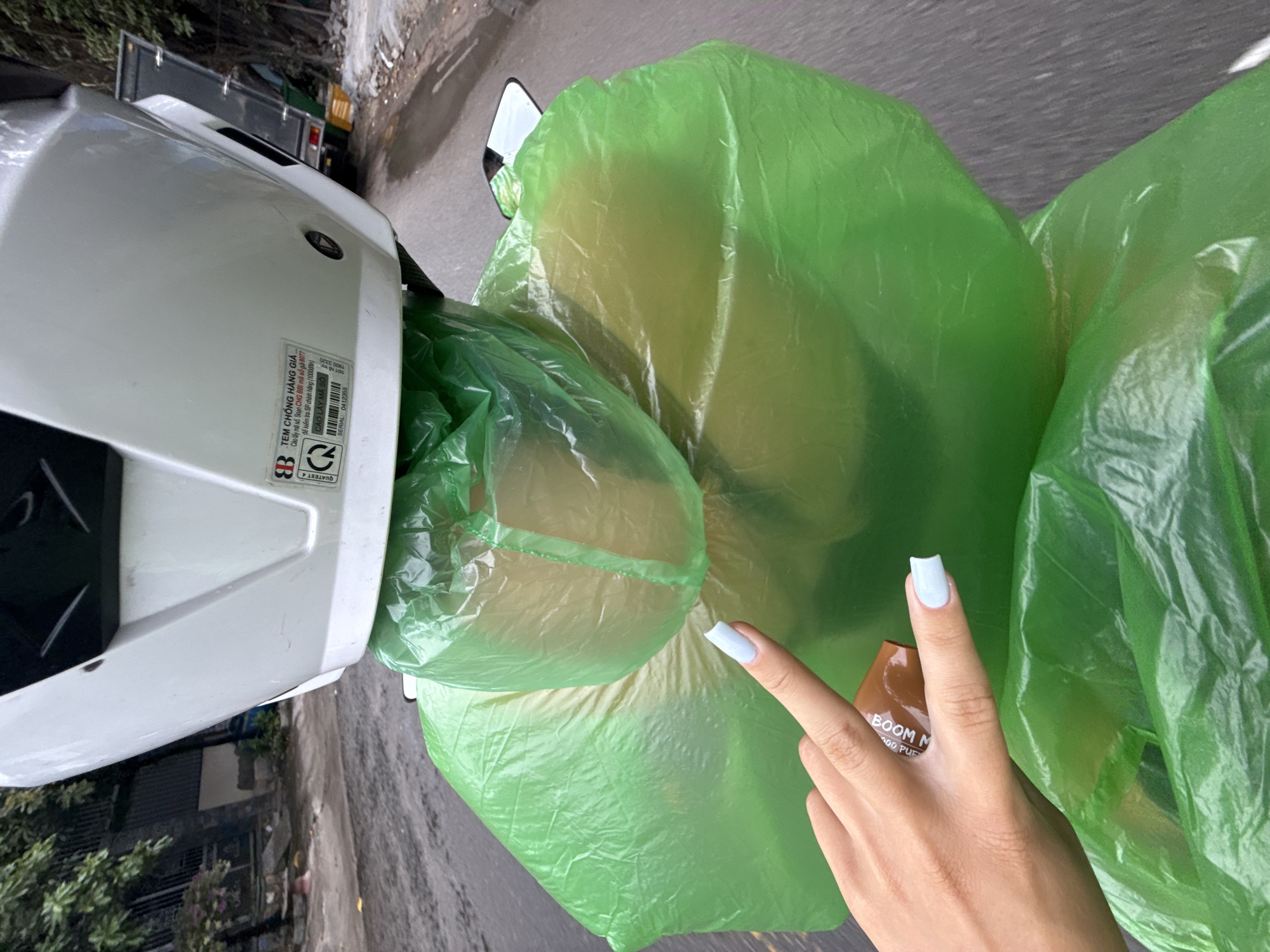 In Nha Trang, Vietnam, a person wearing a white helmet and a bright green poncho is riding a motorcycle. The poncho covers their entire body, obscuring their features. A hand with light blue nail polish is visible, with fingers extended towards the poncho. The person is on a wet, paved road, suggesting it has recently rained or is currently raining. The background shows a fence and some trees, typical of a city environment. The scene appears to be captured from a first-person perspective, likely from the passenger's or rider's point of view.