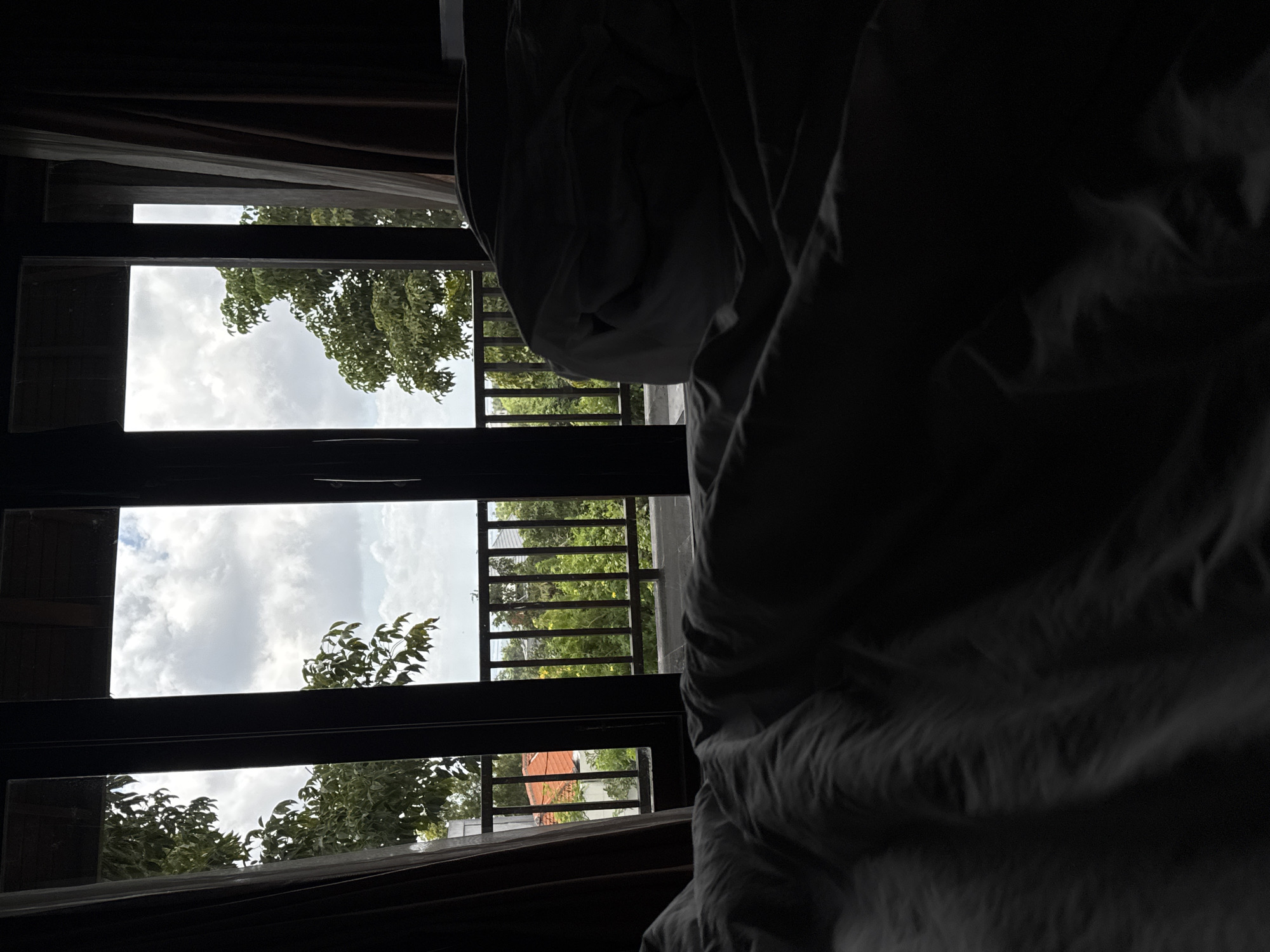 The image, taken from indoors, depicts a view through a window in Kuta Selatan, Indonesia. The window is divided into three horizontal sections, revealing a partly cloudy sky and foliage outside. A balcony railing is visible between the windows. To the right, a white sheet or fabric is prominently featured, occupying a significant portion of the frame. The indoor setting is dimly lit, creating a contrast with the brighter outdoor scene. The image is taken in a residential setting.