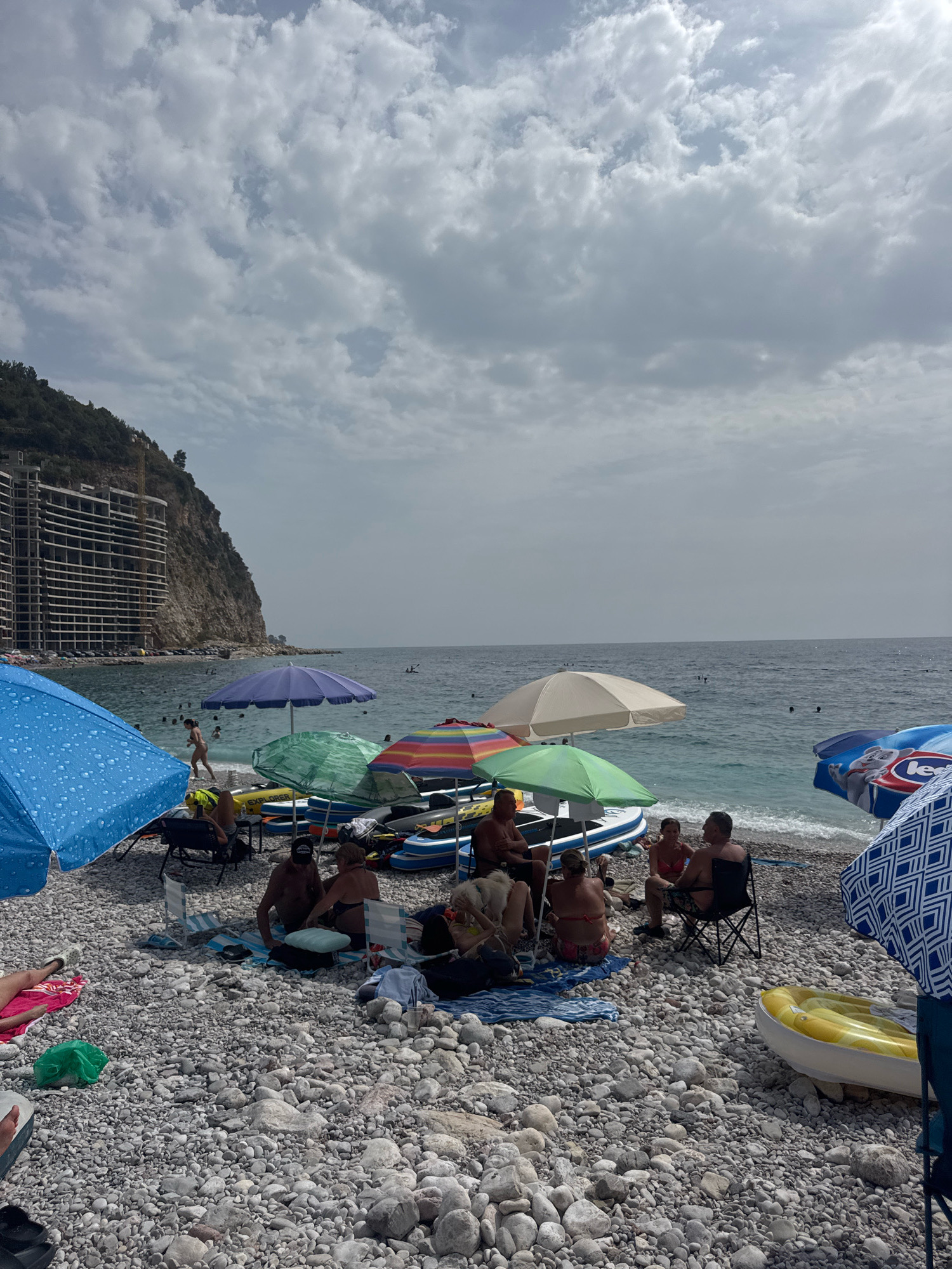 This is a photograph of a crowded beach scene on a cloudy day. In the foreground, several beach umbrellas are set up, providing shade for groups of people. These groups are engaged in various activities, including sitting in chairs, conversing, and lounging on towels. Various inflatable watercraft, such as kayaks and dinghies, are visible behind the umbrellas. The beach itself is covered in pebbles. In the background, the sea is visible with some people swimming or floating in the water. To the left of the frame, a large, unfinished building complex is under construction on a rocky hillside that is covered in trees. The horizon is dominated by a cloudy sky. The overall impression is that of a popular tourist destination.
