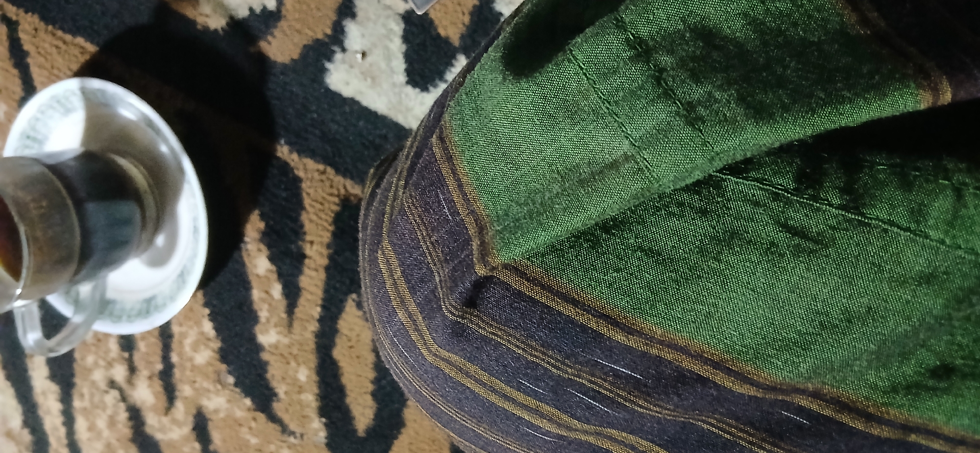 A high-angle, close-up shot captures a striped fabric and a coffee cup on a patterned rug. The fabric, primarily green with dark stripes and thin gold and white pinstripes, is folded, revealing its textured weave. To the right, a clear glass cup filled with dark liquid rests on a white saucer with a green decorative rim. The saucer is placed on a rug with a brown and black tiger-like pattern. The lighting suggests an indoor setting, possibly a domestic environment in Demangan, Indonesia. No people are visible in the frame.