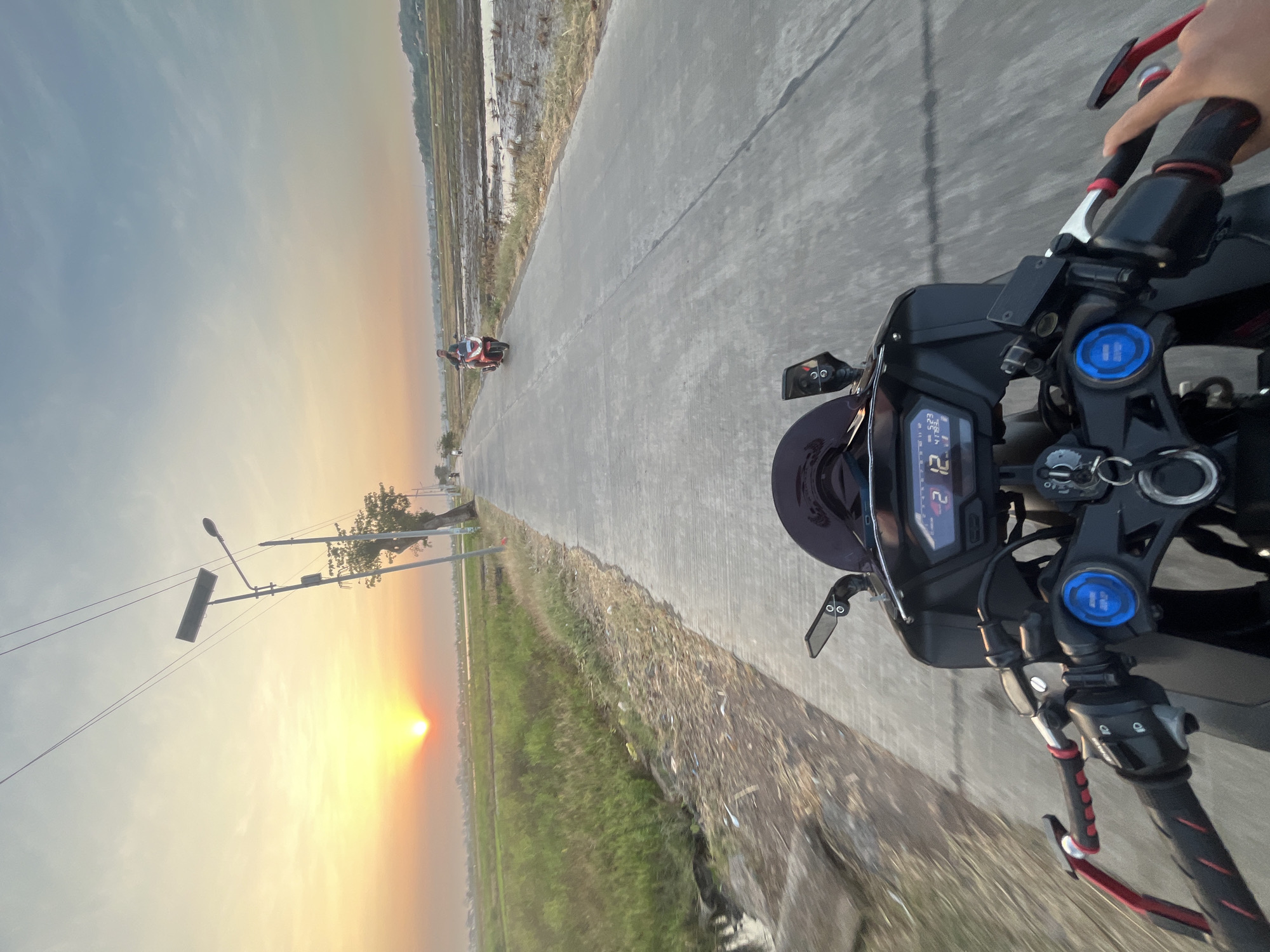 The image is a first-person perspective shot from a motorcycle on a paved road in Pecangaan, Jepara, Indonesia, during sunset. The sun is low on the horizon, casting a warm, orange glow across the sky and illuminating the fields on either side of the road. On the left, there are green fields, and on the right, there are rice paddies with some muddy water. A street light and a solar panel on a pole are visible along the left side of the road, with power lines stretching across the sky. Two other motorcycles are visible in the distance on the road. The motorcycle in the foreground has a digital speedometer displaying "12 5:23 41753". The handlebars and front of the motorcycle are in focus. The overall scene depicts a serene rural landscape at dusk.