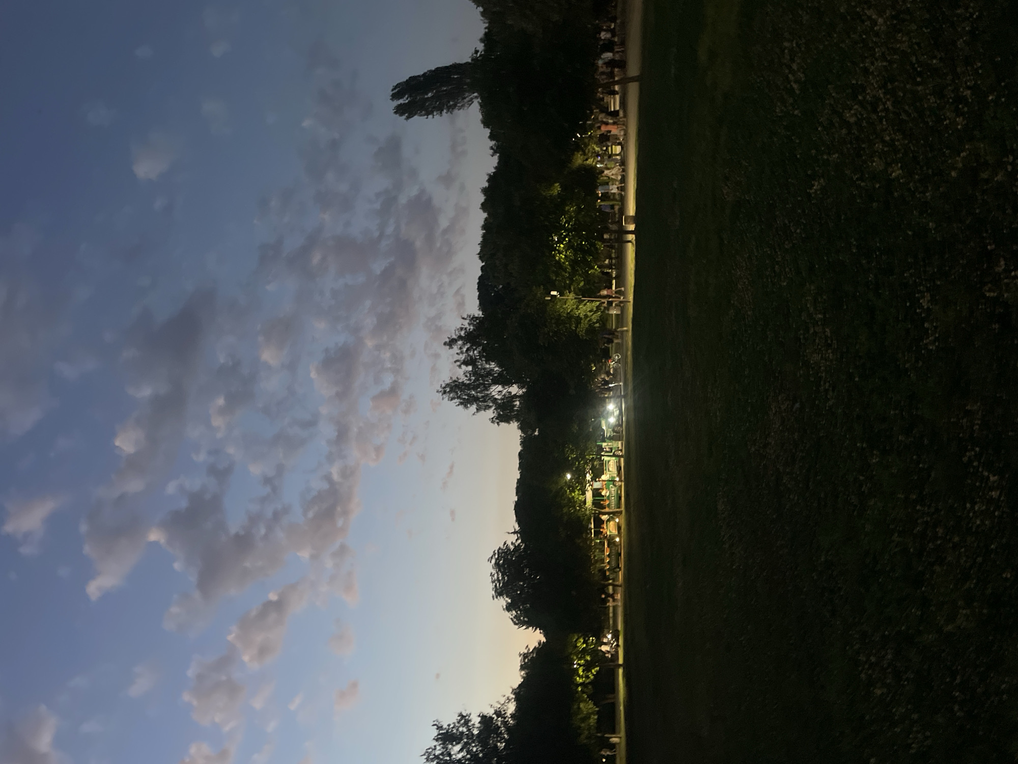 The image depicts a twilight scene in Burgas, Bulgaria, likely in a park or recreational area, given the presence of trees, open grassy areas, and pathways. In the background, a row of trees is illuminated by artificial lights, revealing several people and a green tourist train labeled "GREEN LIFE." The train appears to be stopped or moving slowly, with passengers visible. Further back, more trees form a dark canopy. The sky is a deep blue with scattered, wispy clouds, indicating the transition from dusk to night. The foreground is a dark, grassy field with some scattered fallen leaves or debris. The overall atmosphere suggests an evening outing or event in a public space.