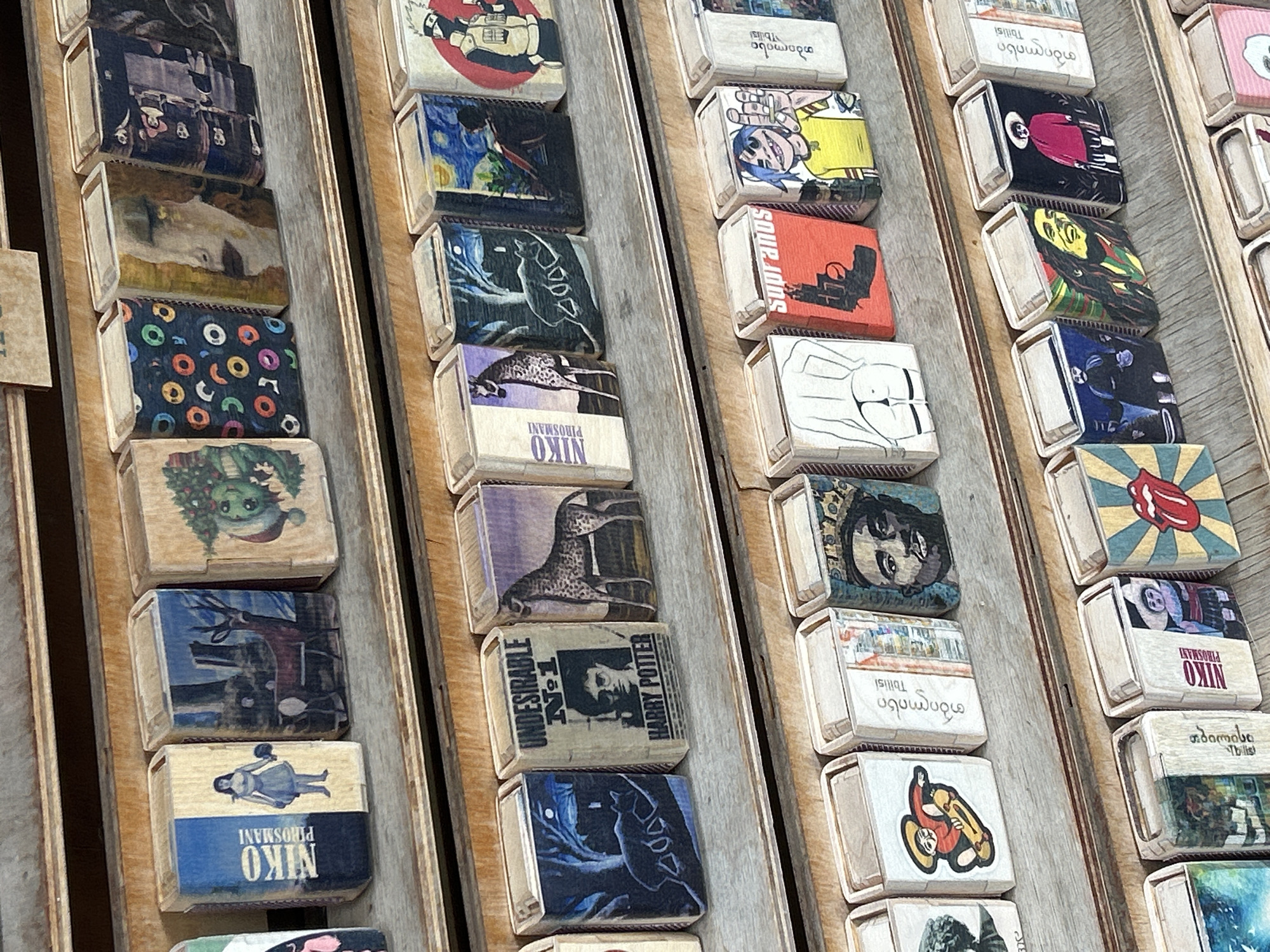 The image shows a close-up, eye-level view of several shelves of decorative matchboxes. The matchboxes are arranged in rows and appear to be made of wood. Each matchbox is adorned with various colorful illustrations. The subjects of the illustrations include portraits of people, animals, abstract patterns, and pop culture figures. The shelves themselves are made of wood and are part of a display, likely in a shop. The overall impression is one of a curated collection of artistic matchboxes. The context provided suggests this scene is located in Mtskheta, Georgia.