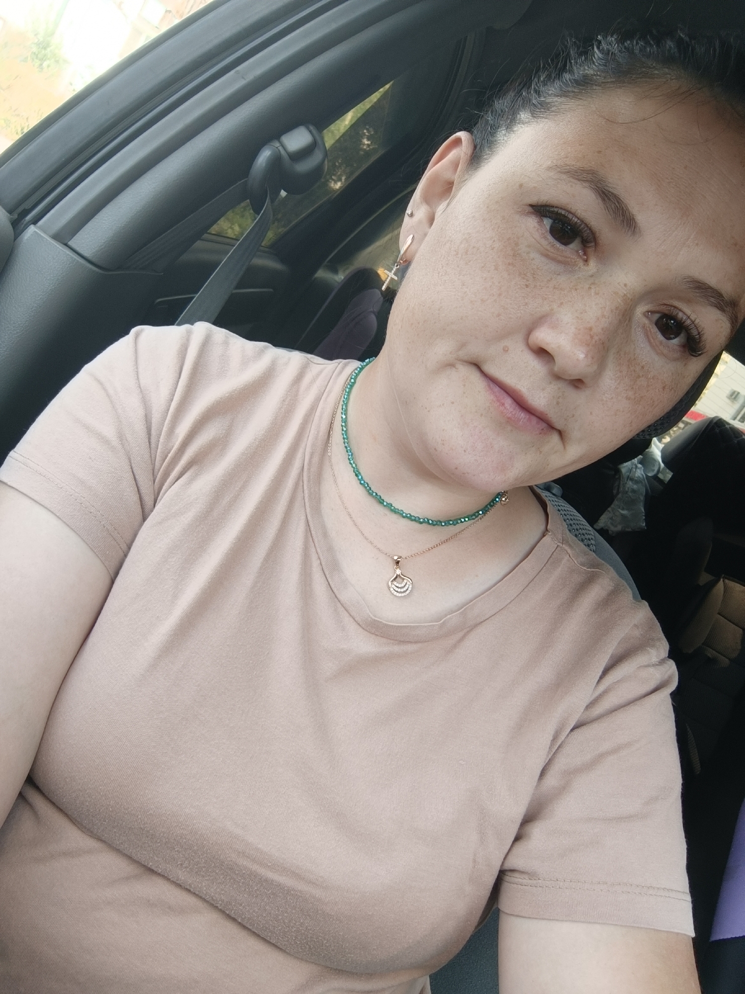 The image shows a close-up, selfie-style shot of a woman sitting in the driver's seat of a car. She has dark hair pulled back, prominent freckles on her face, and is wearing makeup including eyeliner and false eyelashes. She has a green beaded necklace and a delicate rose gold necklace with a pendant. She is wearing a plain, light brown t-shirt. The interior of the car is visible, including the seatbelt, car door panel, and part of the headrest. Sunlight is streaming in from outside, suggesting it is daytime. The woman's expression is neutral. There is no visible text in the image. The location context provided is Chaikovsky, Russia.
